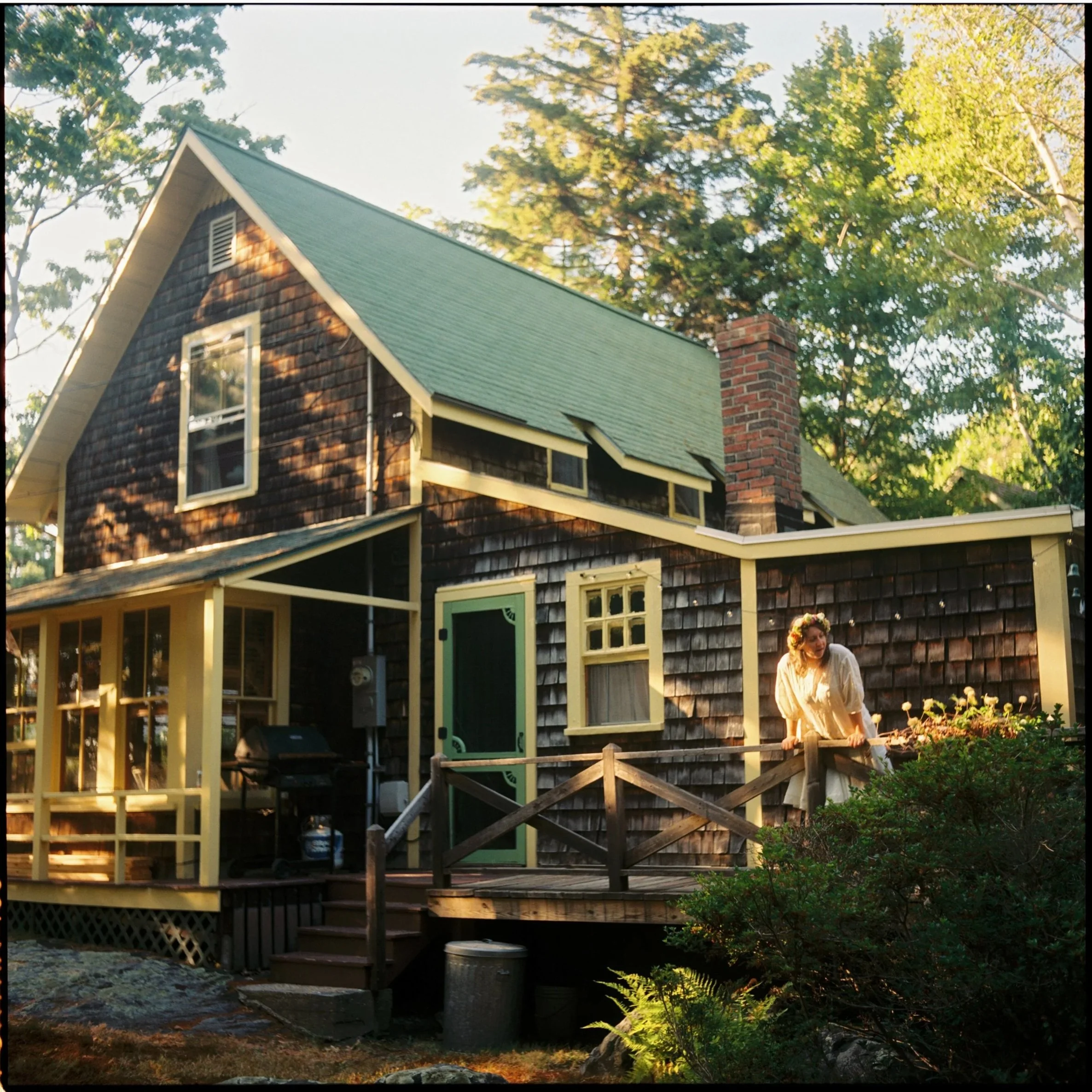 A woman with a flower crown looking over a wooden deck at a rustic house with dark shingles and a green roof, surrounded by trees. Non -traditional film wedding photographer in Maine. 