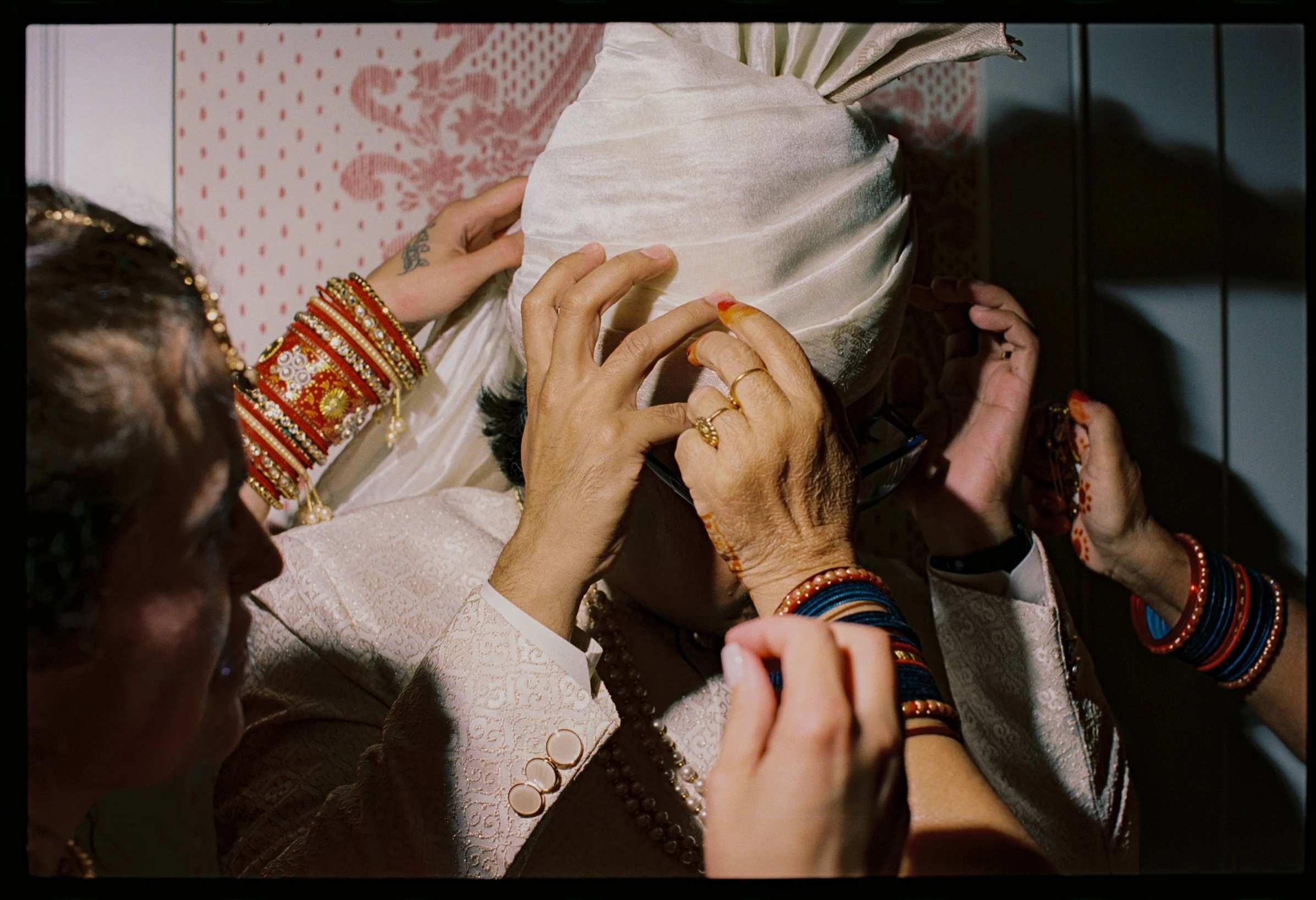 Close-up of hands adjusting a cream-colored traditional Indian wedding attire, decorated with jewelry and vibrant bangles, against a pink patterned background. Non -traditional film wedding photographer in Maine. 