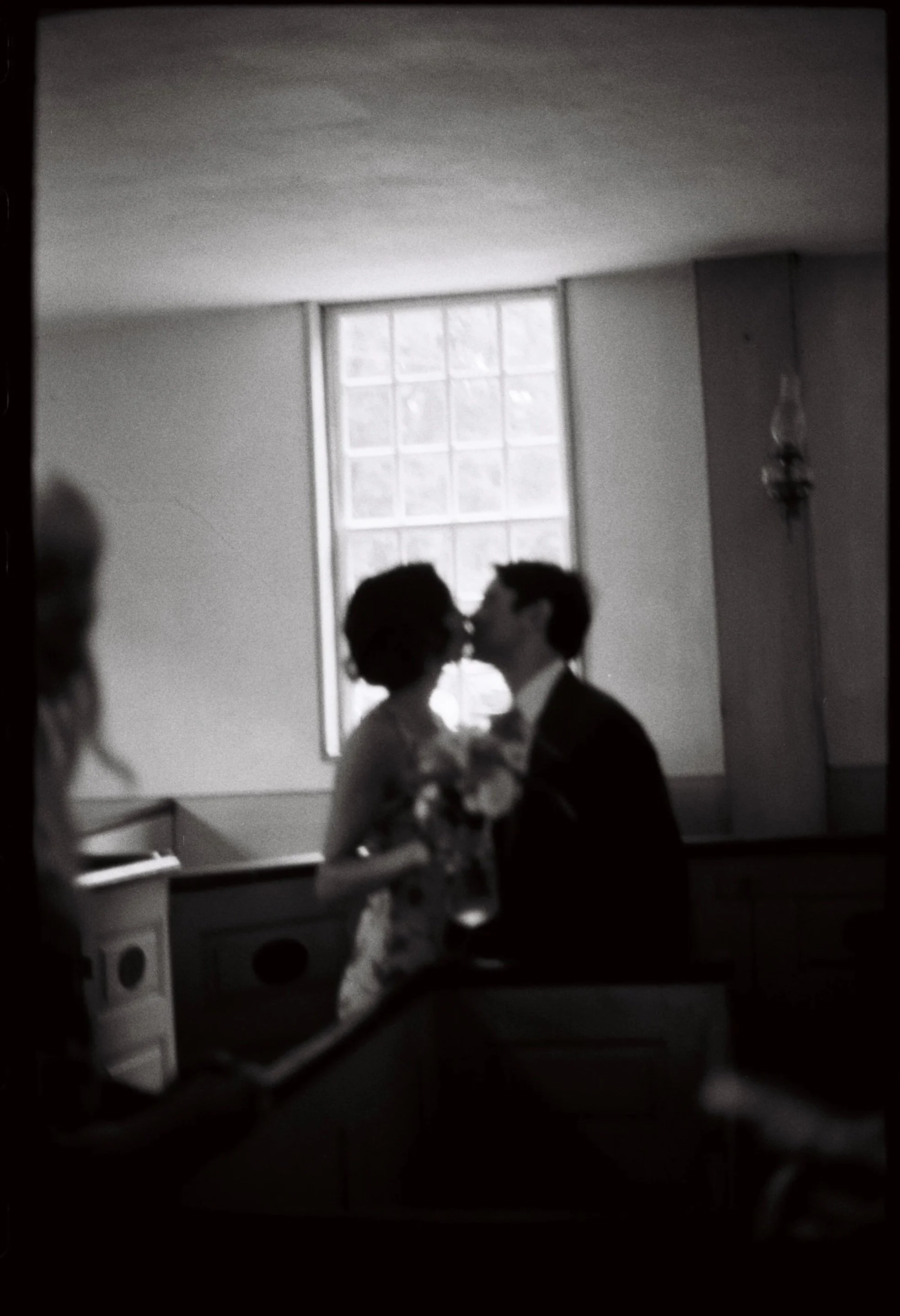 Silhouette of a couple kissing, with the woman holding a bouquet of flowers, inside a dimly lit room with a large window. Non -traditional film wedding photographer in Maine. 