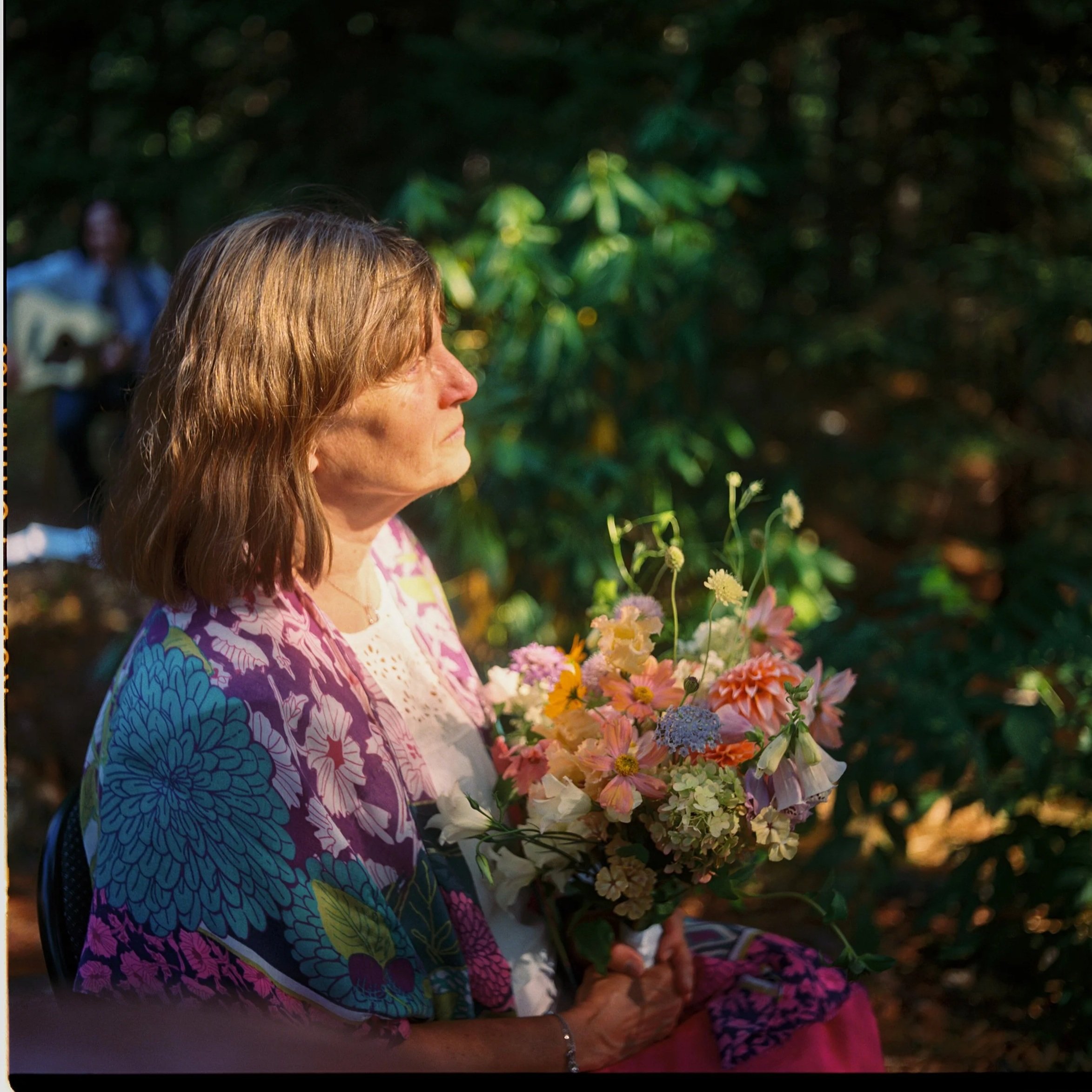 A woman with light brown hair sitting outdoors holding a bouquet of colorful flowers, wearing a floral patterned shawl, with green foliage in the background. Non -traditional film wedding photographer in Maine. 