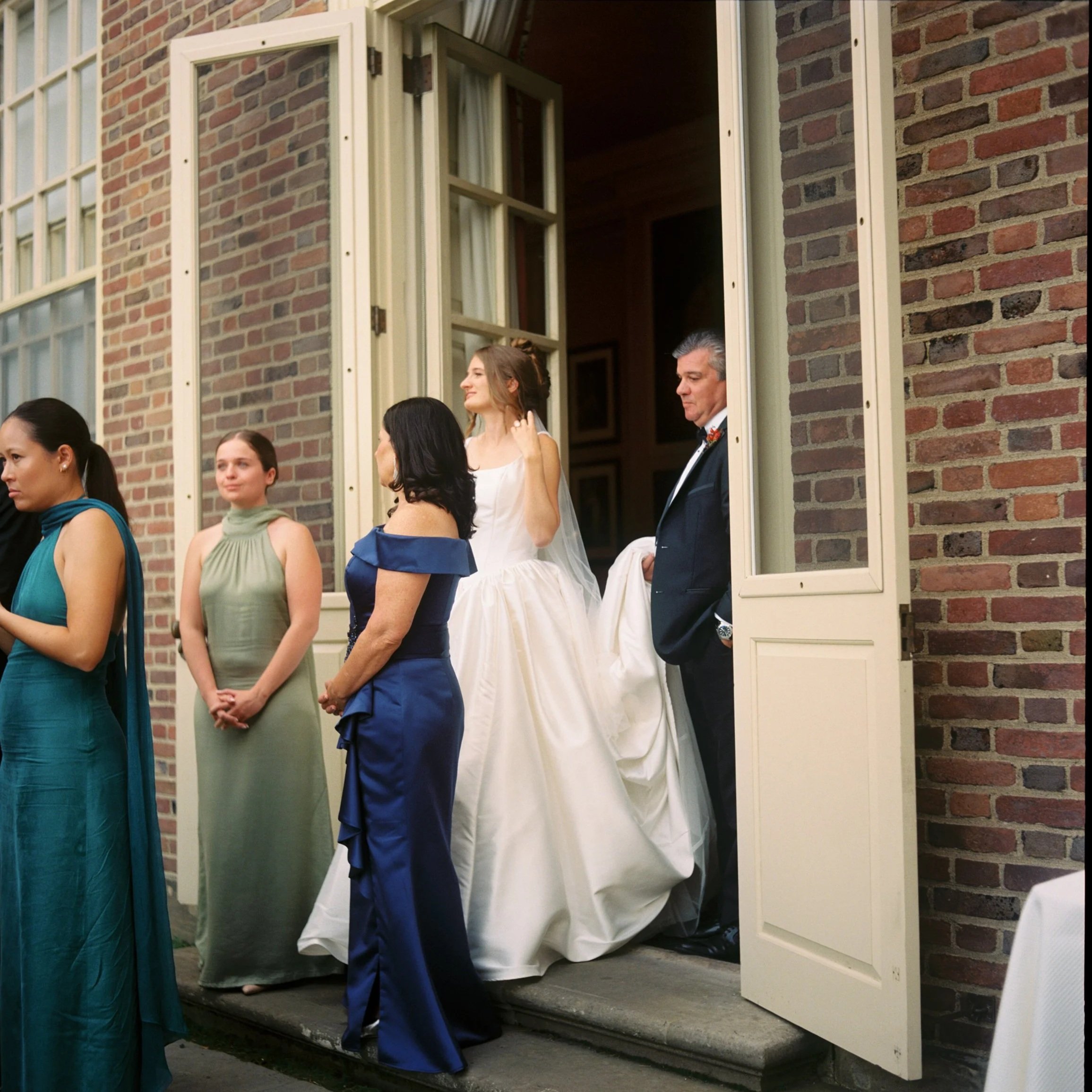 Bride and groom with guests standing outside near open window during wedding ceremony in a brick building. Non -traditional film wedding photographer in Maine. 