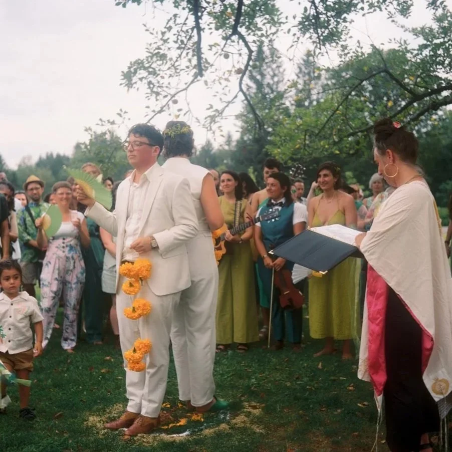 A wedding ceremony outdoors with a couple dressed in white, decorated with marigold flowers. The groom wears glasses and holds a fan, while the bride, with her back to the camera, wears flowers in her hair. Guests surround them, some holding fans and