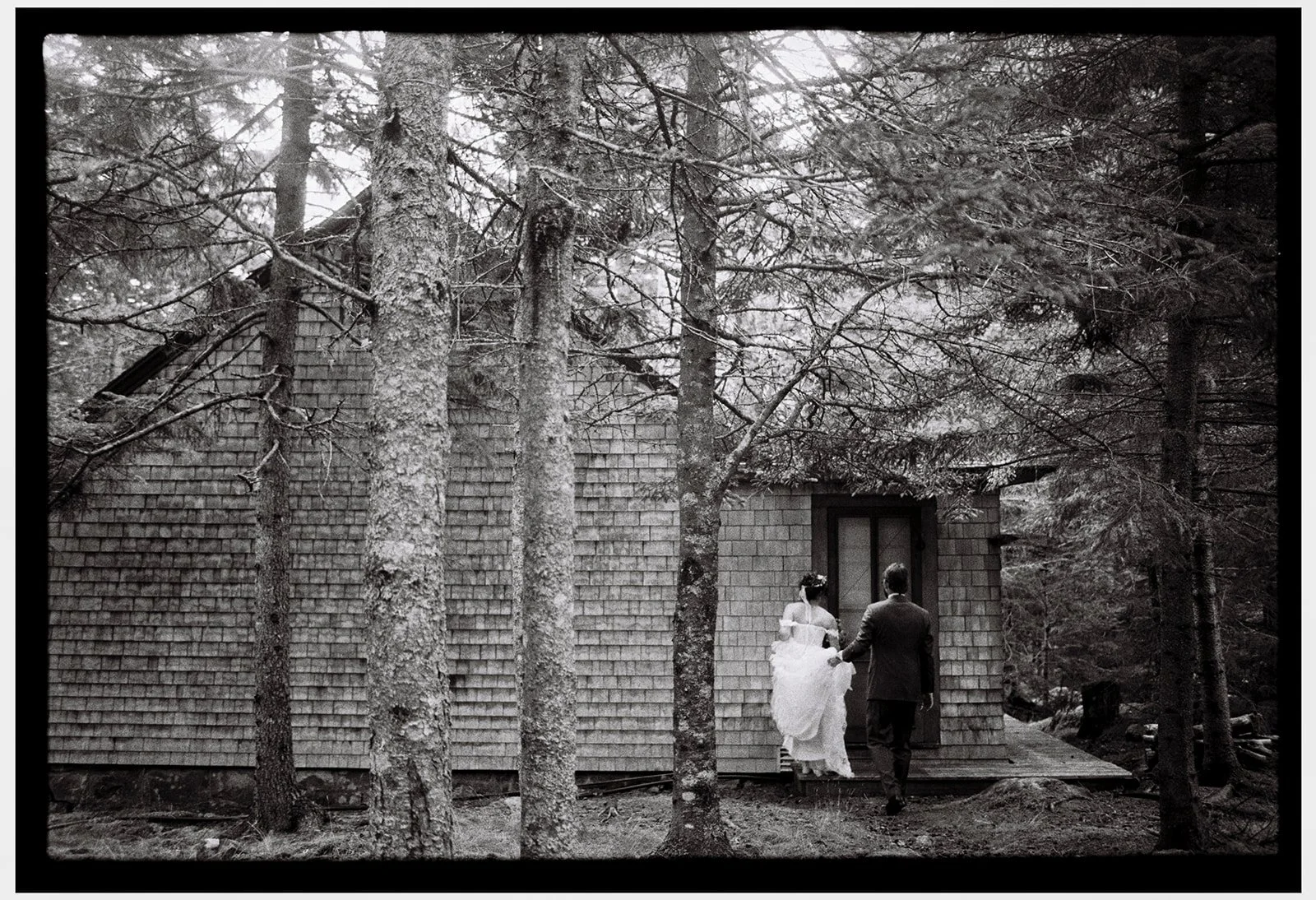 A black and white photo of a couple walking towards a house in a forest, with tall trees surrounding the building. Non -traditional film wedding photographer in Maine. 