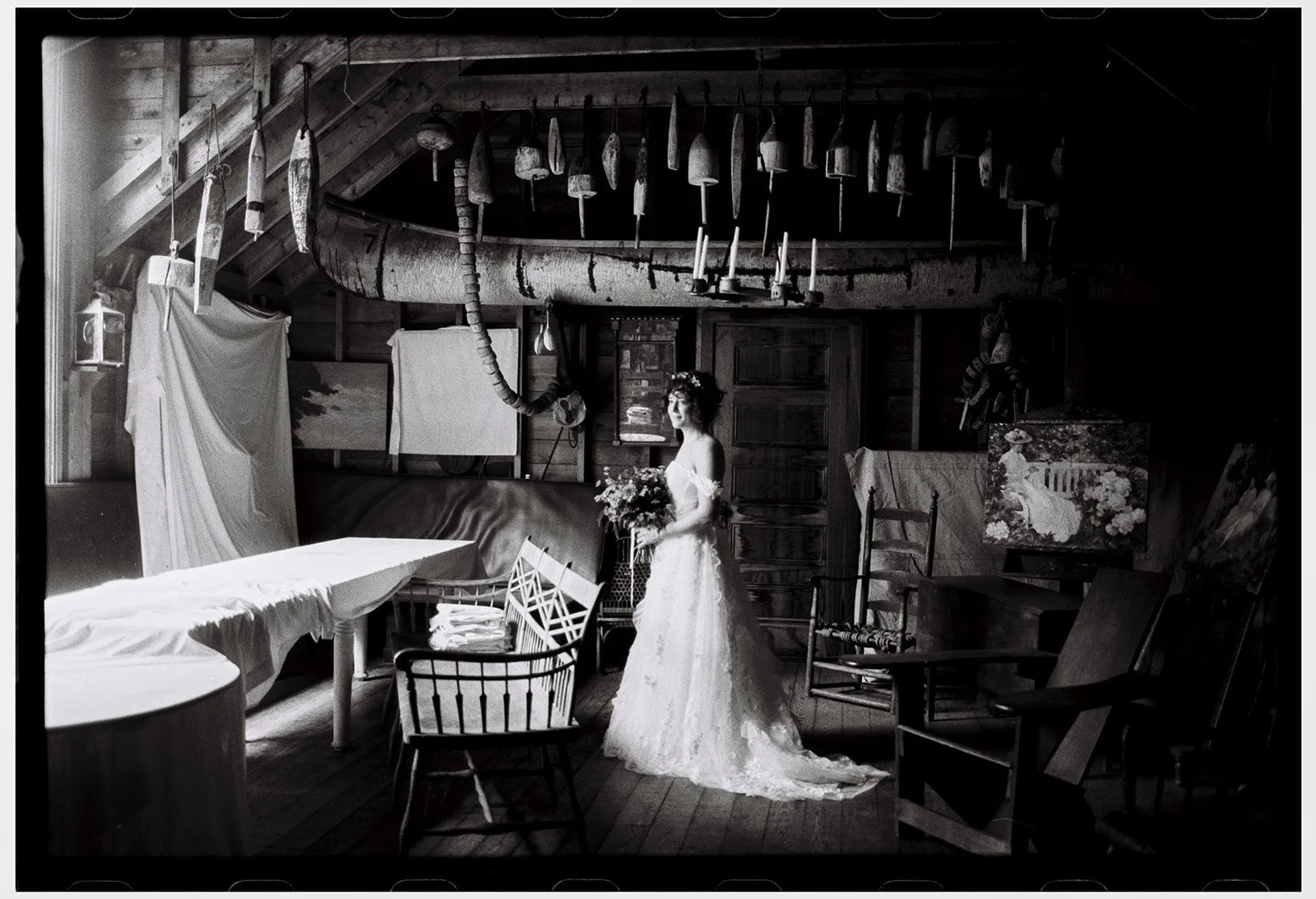Black and white photo of a woman in a wedding dress holding a bouquet in a rustic room with wooden walls, various hanging tools, artwork, and furniture. Film wedding photographer in Maine. 
