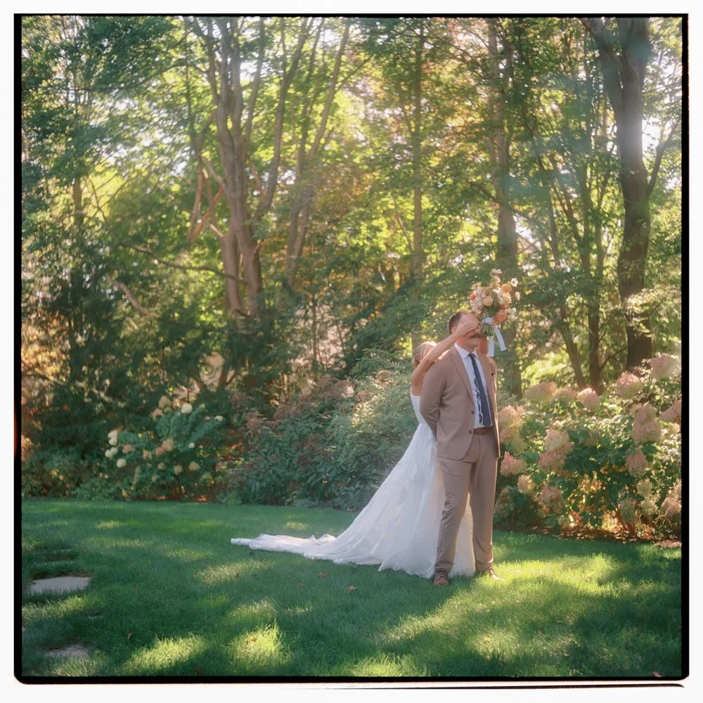 A bride and groom stand together in a lush, green outdoor setting with trees and flowering bushes; the bride partially hides behind the groom as she holds a bouquet over his head. The sunlight filters through the trees, creating a warm, romantic atmo