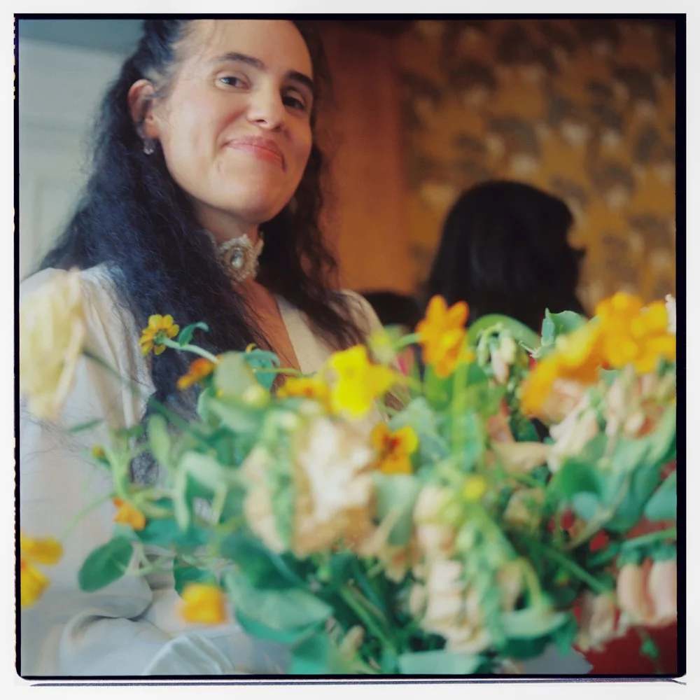 A woman with long dark hair smiling, seated at a table with a floral arrangement of yellow and white flowers in front of her, in a warmly lit indoor setting with a stone wall in the background. Non -traditional film wedding photographer in Maine. 