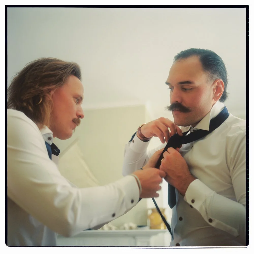 Two men with mustaches wearing white dress shirts and black ties, with one helping the other tie his tie. Non -traditional film wedding photographer in Maine. 