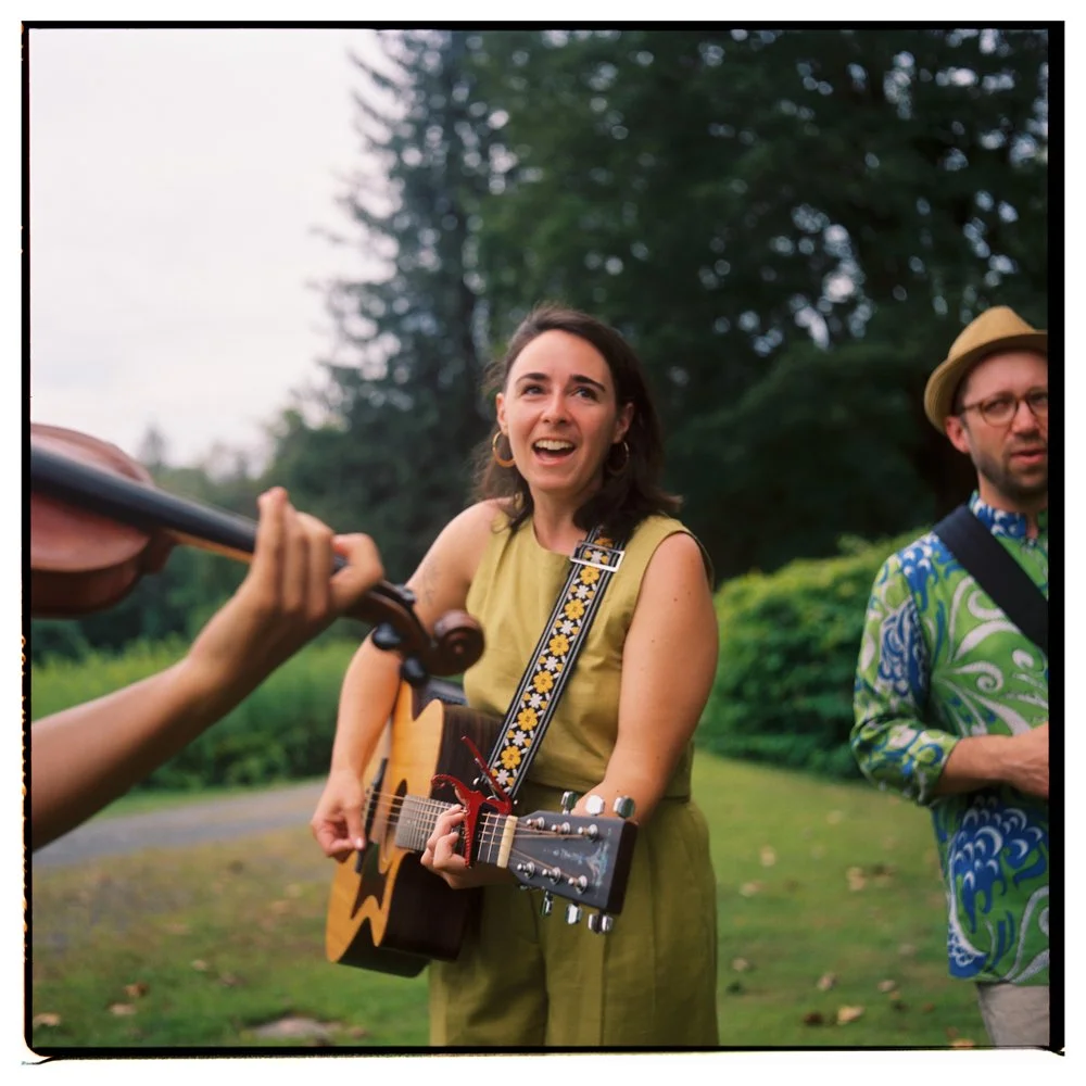 A woman playing an acoustic guitar and singing outdoors as a person with a violin plays nearby, with trees and greenery in the background. Non -traditional film wedding photographer in Maine. 