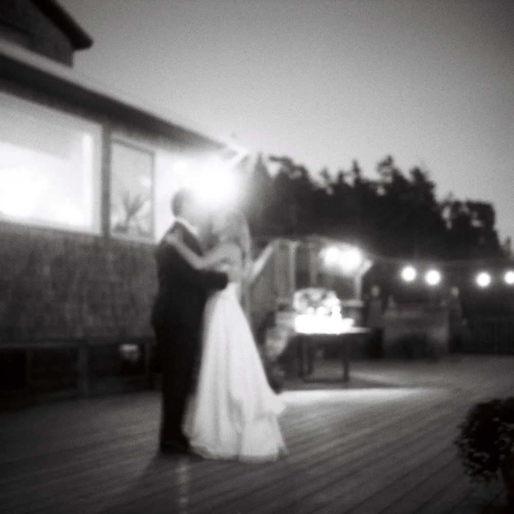 A couple is dancing at an outdoor event in black and white, with party lights and a DJ booth in the background. Non -traditional film wedding photographer in Maine. 