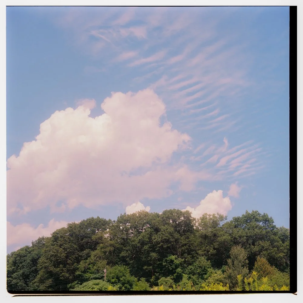 Blue sky with scattered pink-tinged clouds above a green, tree-covered hillside. Non -traditional film wedding photographer in Maine. 