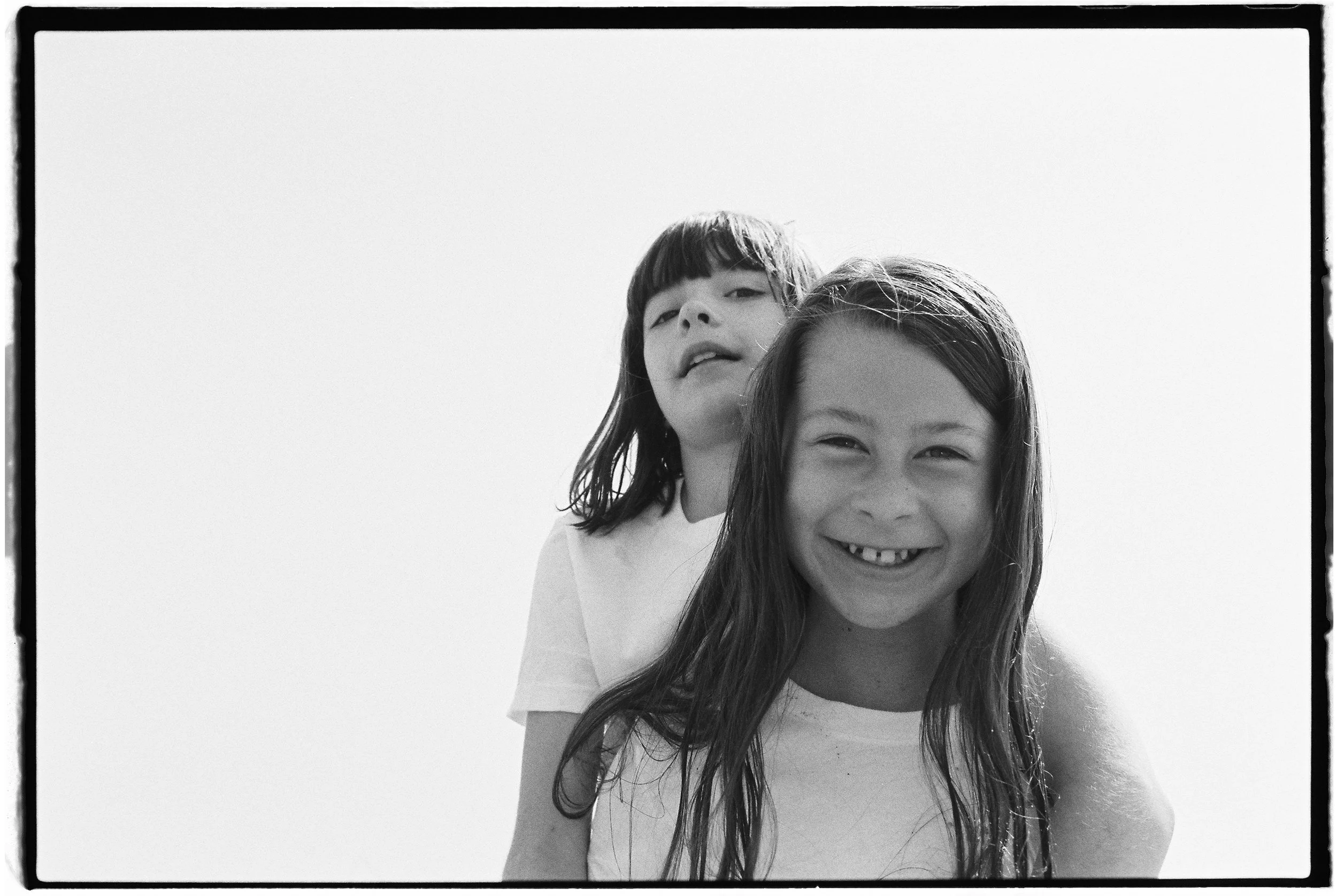 Two young girls, one smiling with missing front teeth and the other with a neutral expression, standing outdoors against a bright sky.