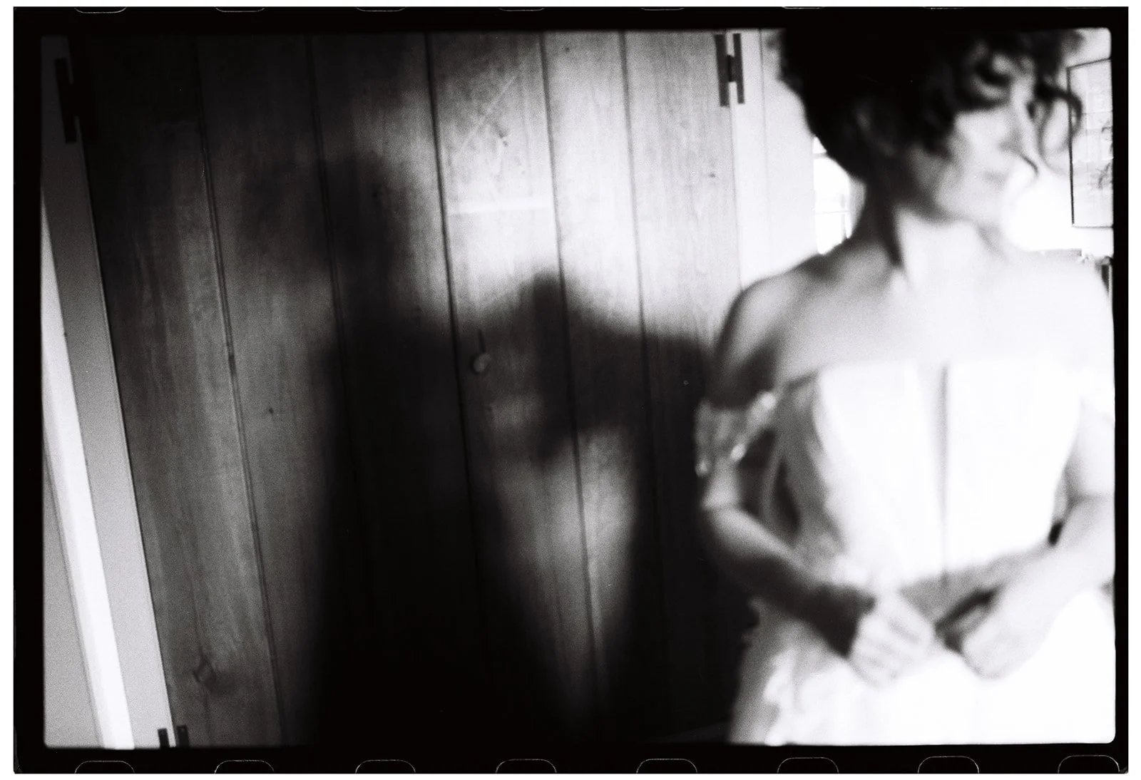 A black and white photo of a woman wearing glasses and an off-the-shoulder dress, standing in front of a wooden wall, with her head turned to the right and her arms crossed.Non -traditional film wedding photographer in Maine. 