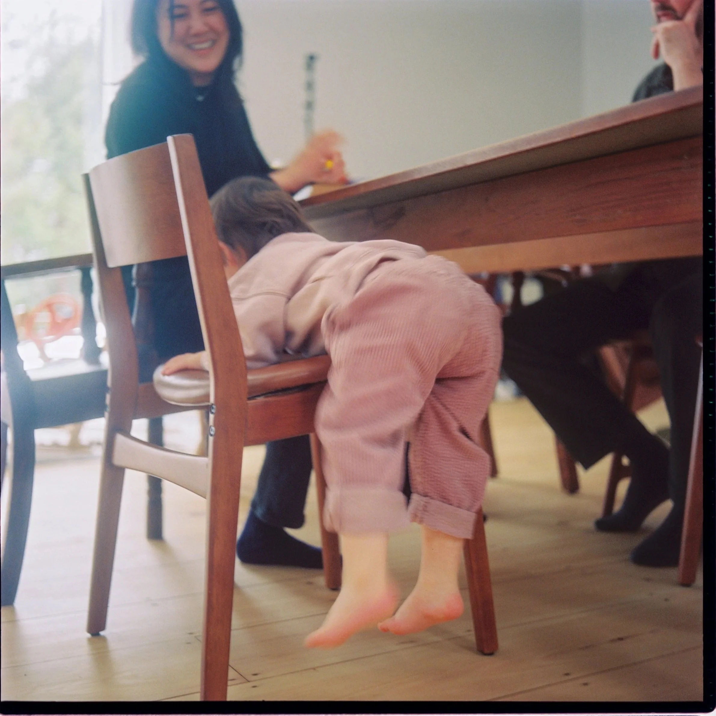 A smiling woman with dark hair sitting at a wooden table, holding a pencil, with a person lying under the table, and a child with bare feet standing nearby, in a bright indoor setting.