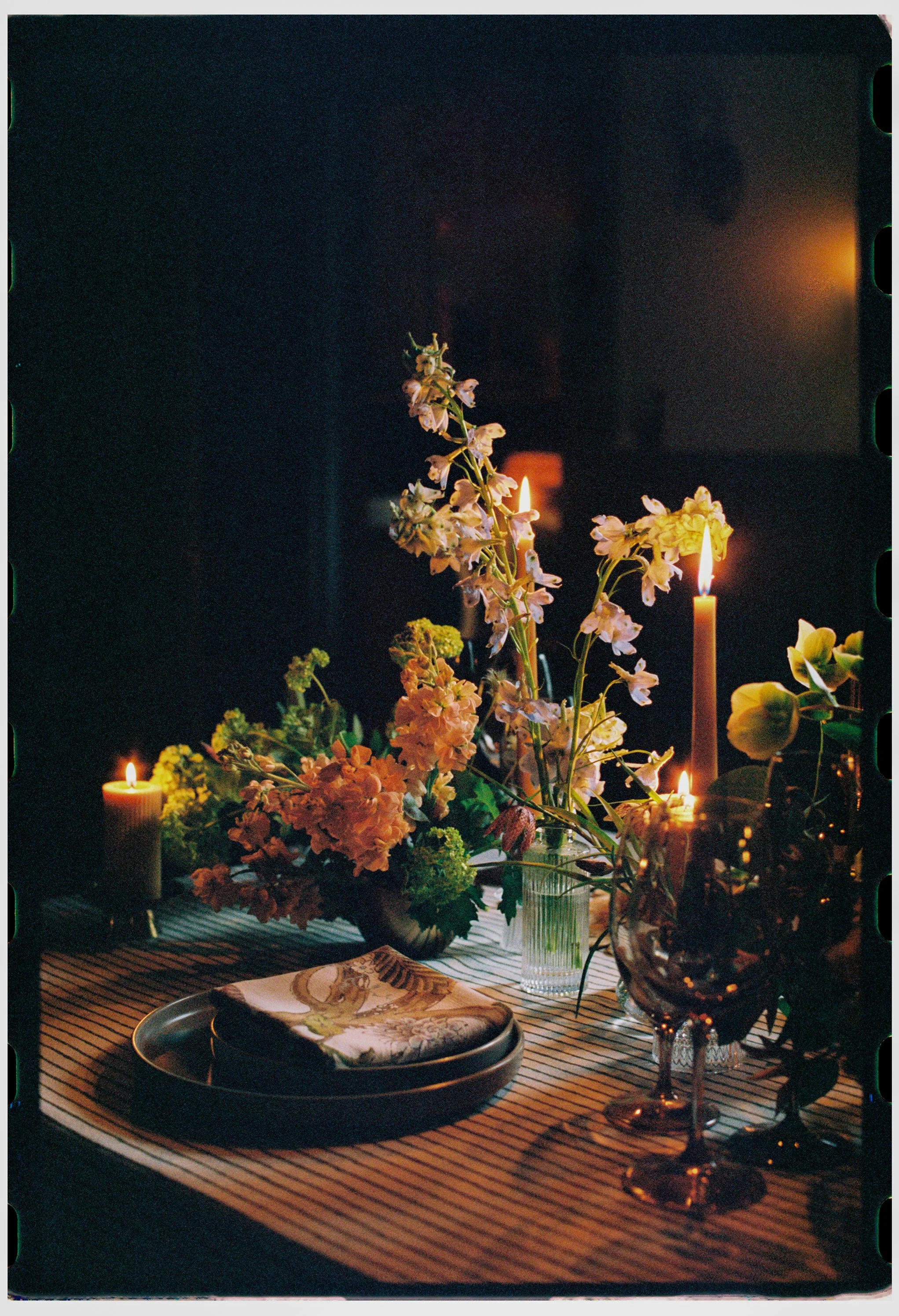 A table decorated with a bouquet of pink and white flowers, lit candles, and a place setting with a napkin and plate in a dimly lit room.