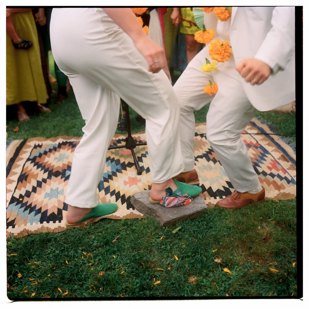 People dressed in white suits participating in a wedding ceremony, with one person stepping on a stone that is part of a traditional sandal dance, wirely decorated with orange flowers, on a colorful patterned rug outdoors. Film wedding photographer. 