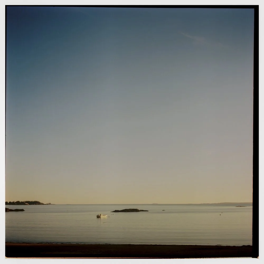 A calm ocean scene at sunset with a small boat floating near rocks in the water and a clear sky above. Non -traditional film wedding photographer in Maine. 