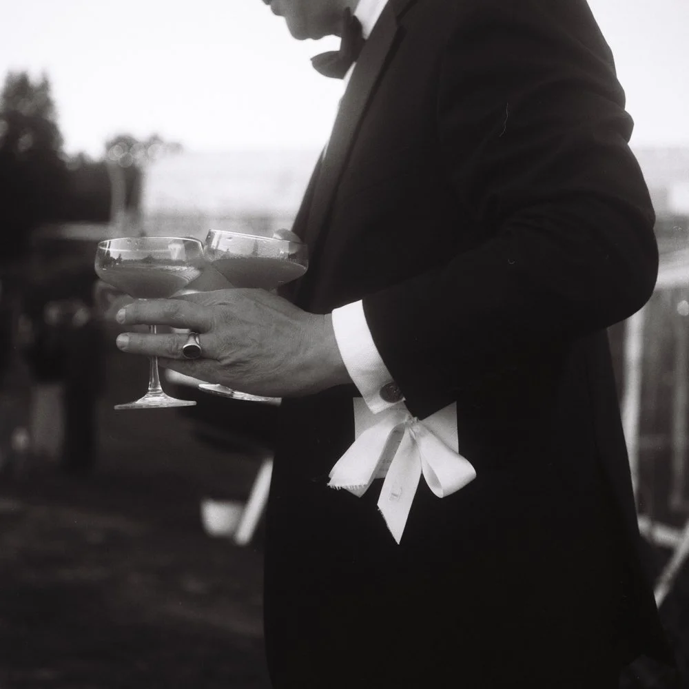 A person in a tuxedo holding several cocktail glasses. Film wedding photographer in Maine. 