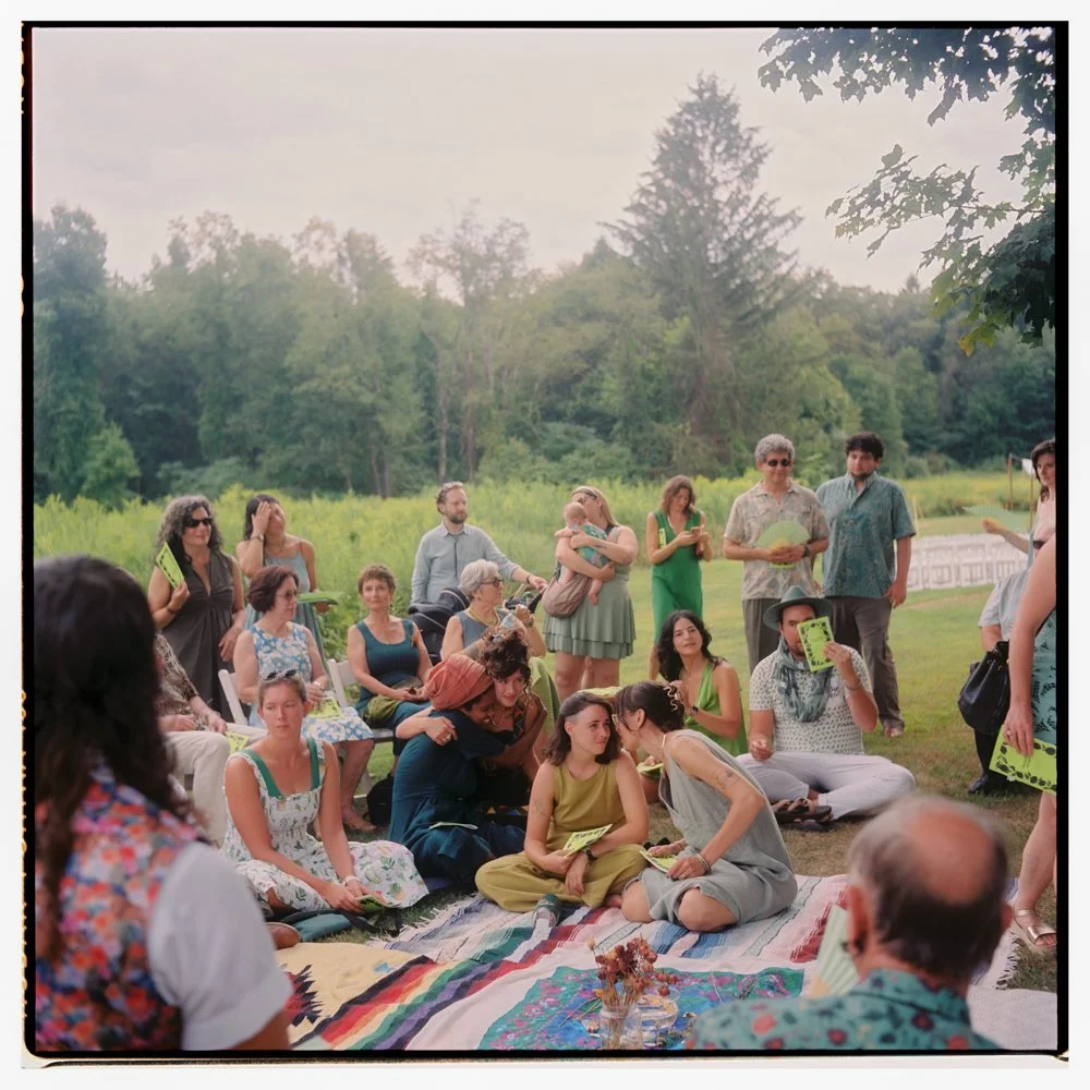 People gathered outdoors in a circle, some sitting on blankets and others standing, engaging in conversation at a picnic or social event with trees and greenery in the background. Non -traditional film wedding photographer in Maine. 