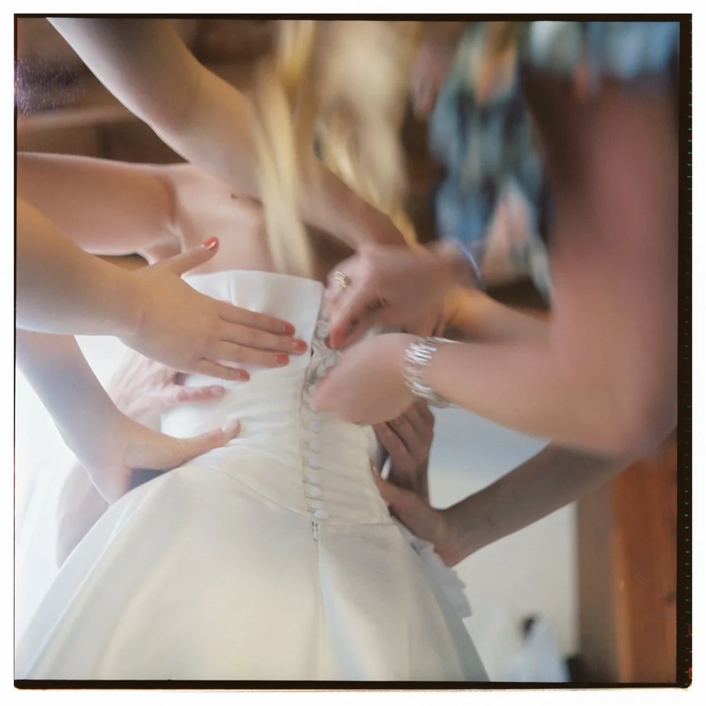Close-up of a bride in a wedding dress being assisted by several women to zip up the back of her gown. Film wedding photographer in Maine. 