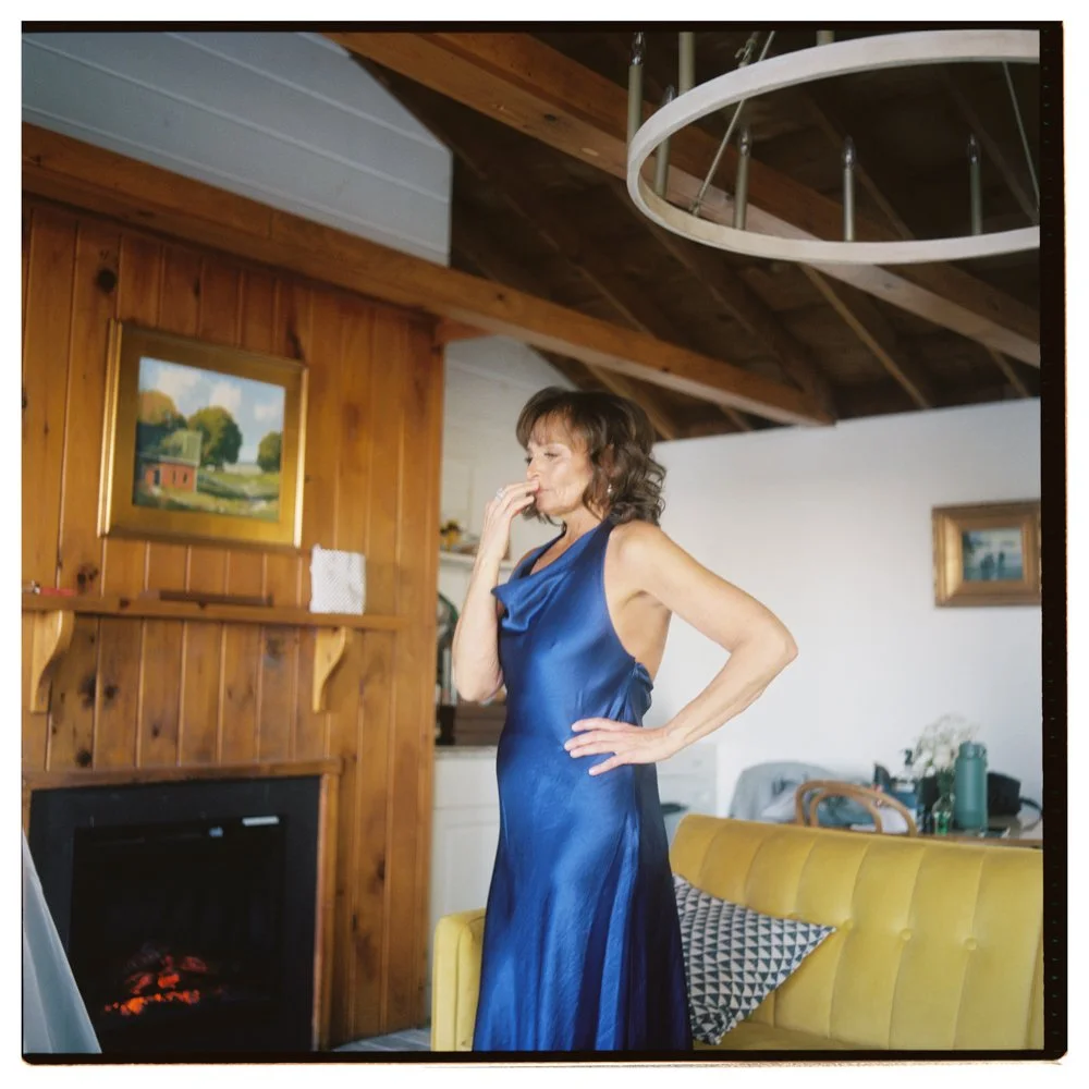 Woman in a shiny blue dress standing in a living room with wood-paneled walls, a yellow couch, and artwork on the walls. Non -traditional film wedding photographer in Maine. 