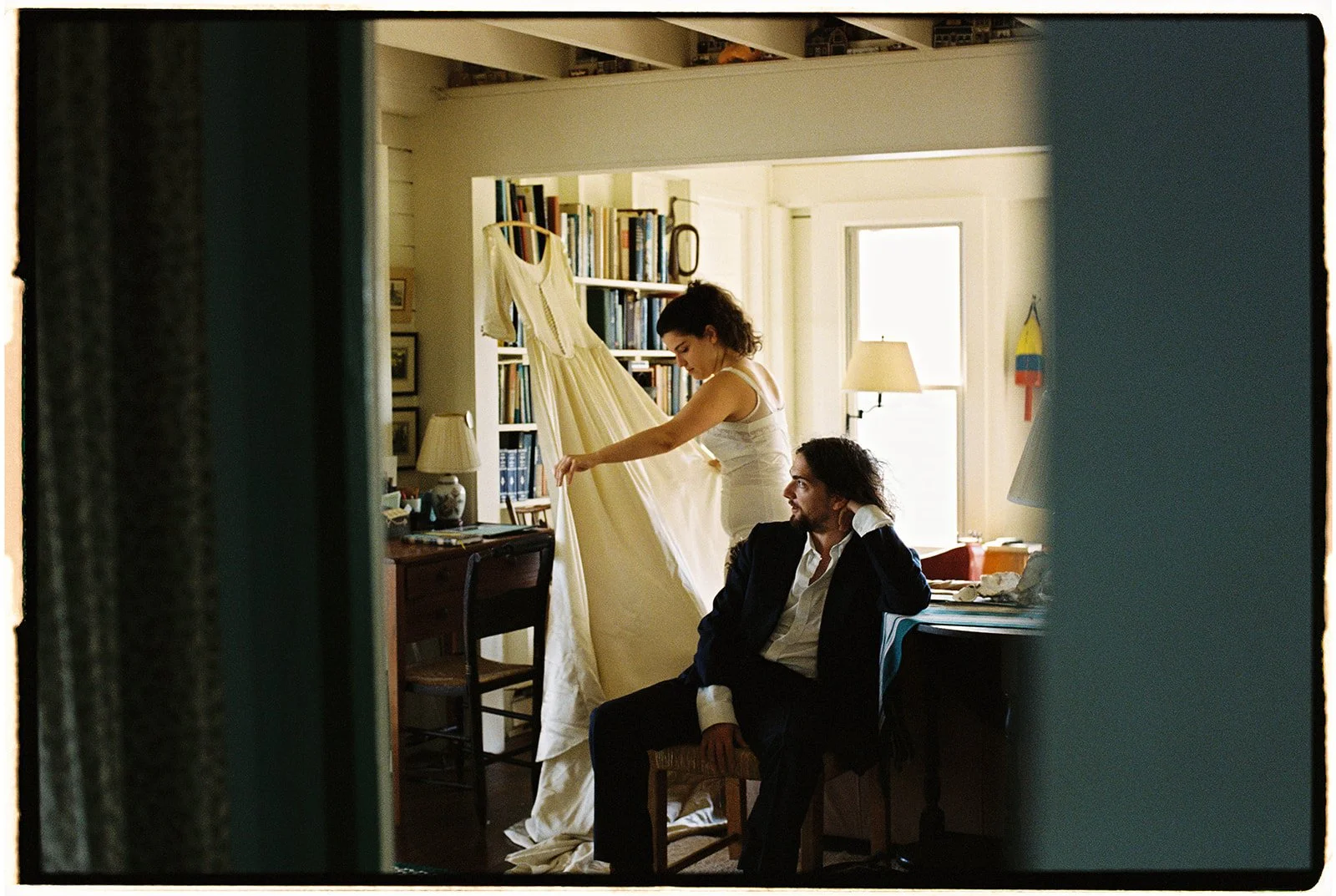 A woman is holding a dress, standing on a chair, next to a seated man inside a cozy room with bookshelves and large windows.