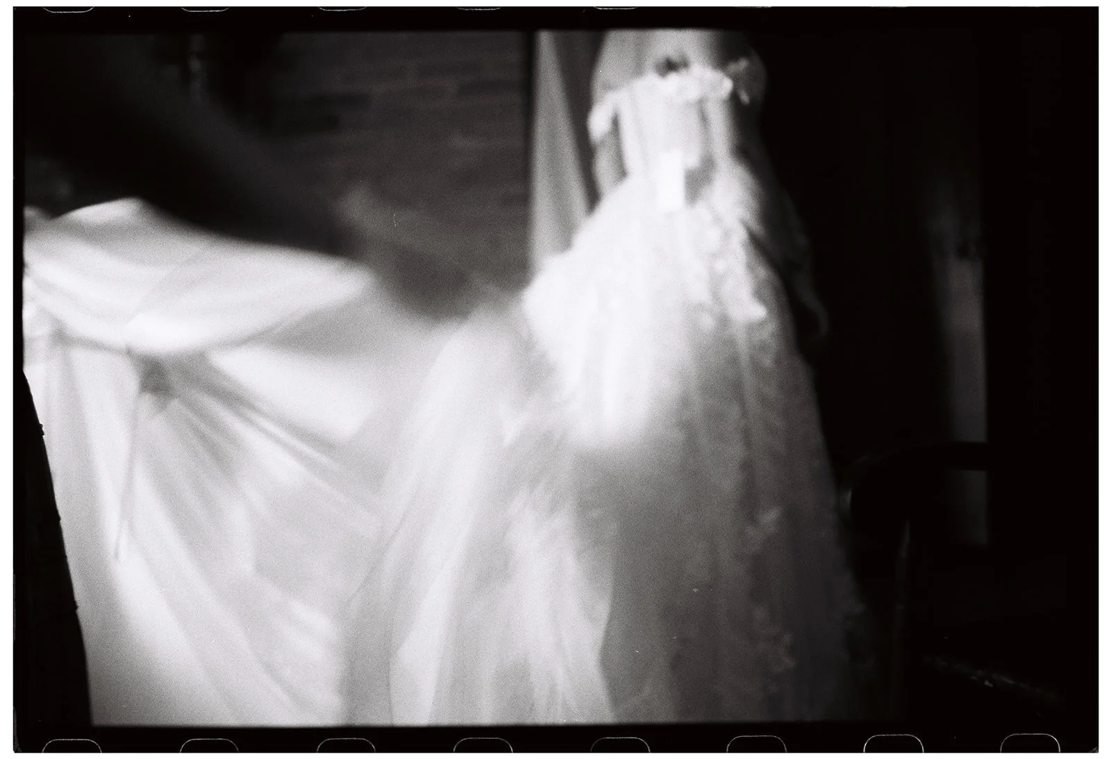 A blurred black and white photo of a wedding dress hanging, with some fabric flowing to the side. Film wedding photographer in Maine. 
