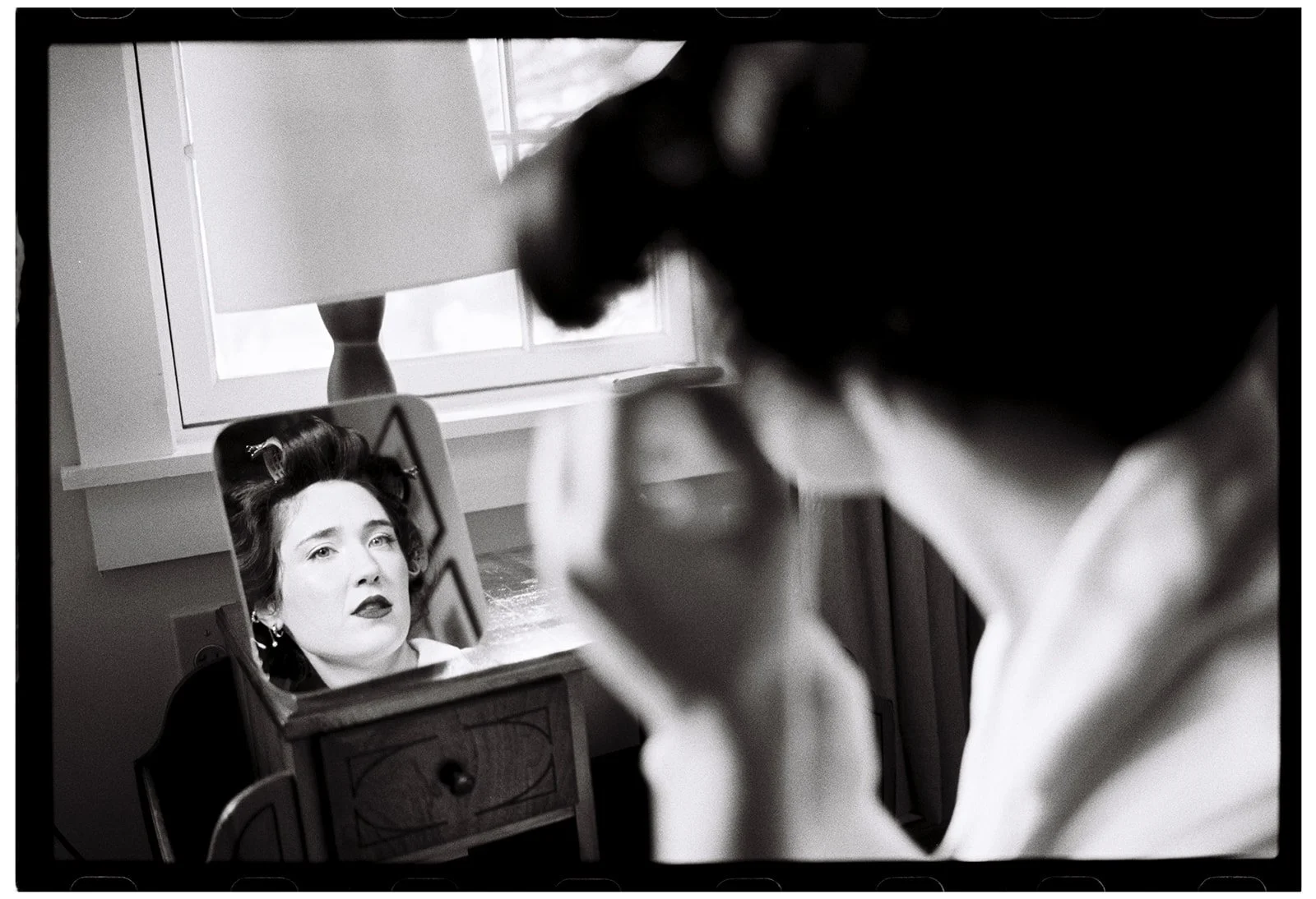 A woman looking at herself in a mirror as a makeup artist applies makeup to her face in a vintage-style setting. Non -traditional film wedding photographer in Maine. 