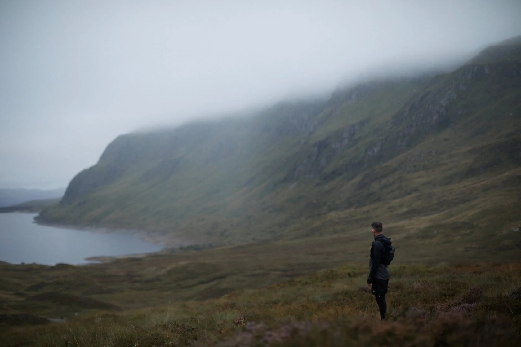 A person in a black jacket and backpack stands on a grassy field near a lake, with misty hills and mountains in the background under cloudy skies.