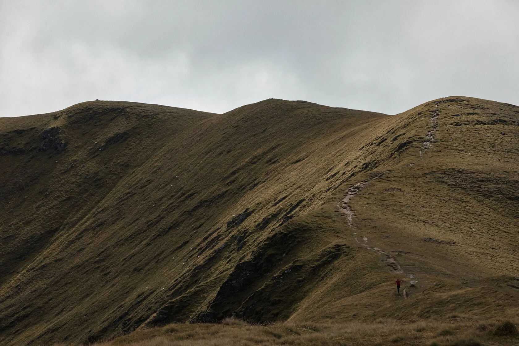 Hilly grassy landscape with a narrow trail winding up a slope, and a person in a red jacket walking along the trail.