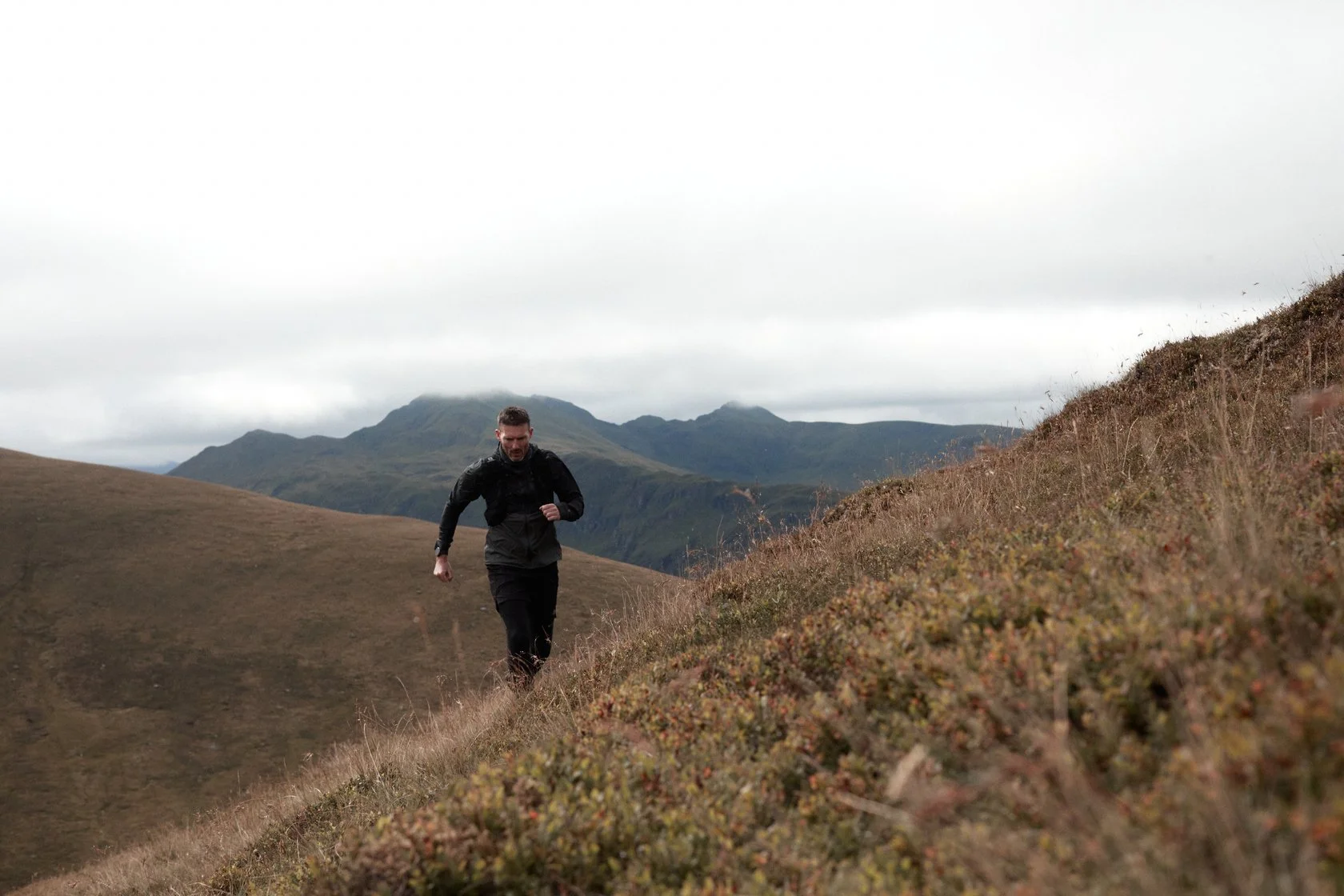 A man dressed in black running uphill through a grassy hilly landscape with mountains and an overcast sky in the background.