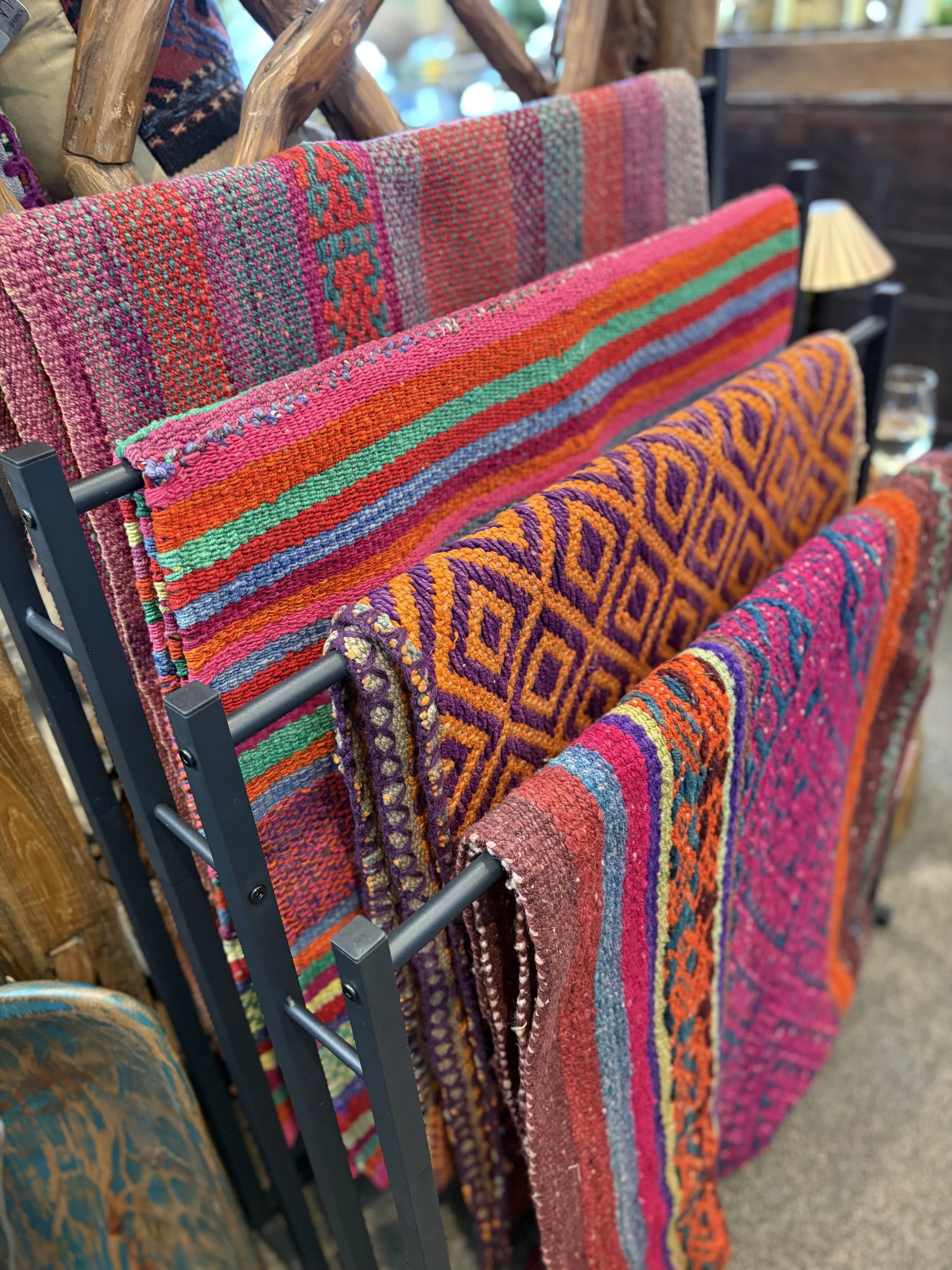 Colorful woven rugs with intricate patterns hanging on a black metal rack, with wooden furniture and a lamp in the background.