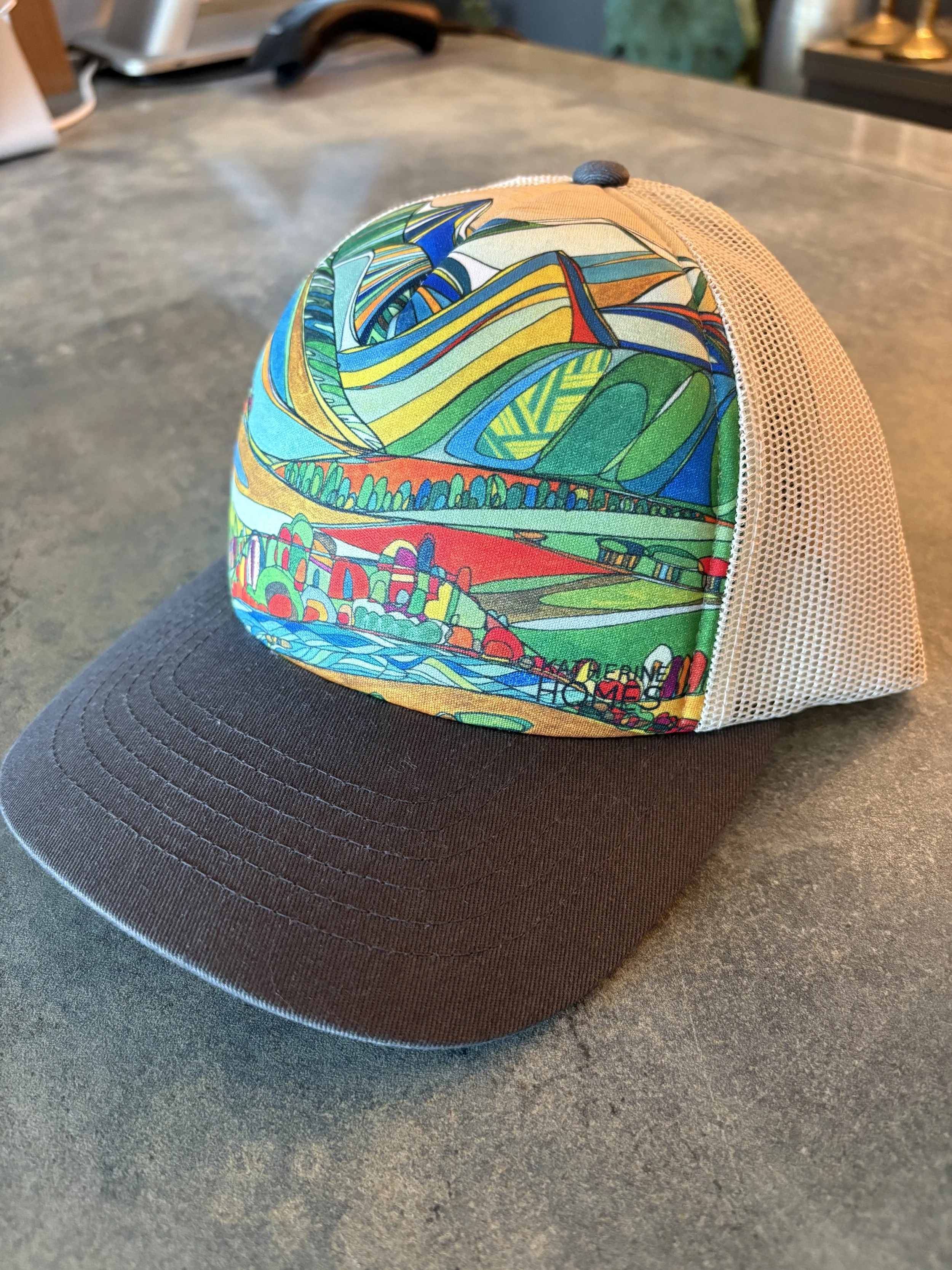 A trucker hat with a brown bill and a mesh back, featuring a colorful abstract design of green, yellow, blue, orange, and red shapes and patterns.
