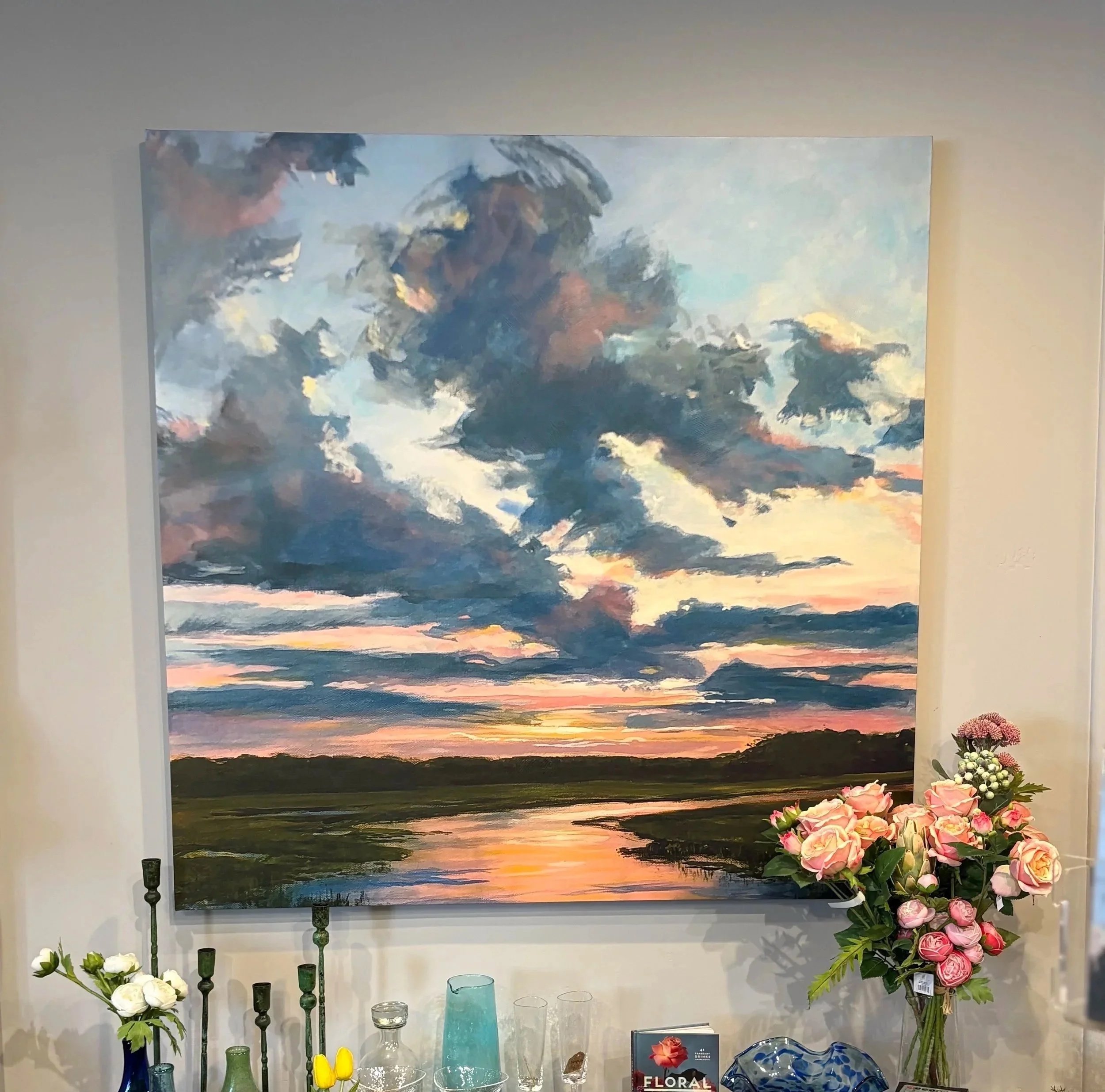 A large landscape oil painting of a sunset sky with dark clouds, pinks, blues, and oranges, hung on a white wall surrounded by decorative flowers and glassware on a table.