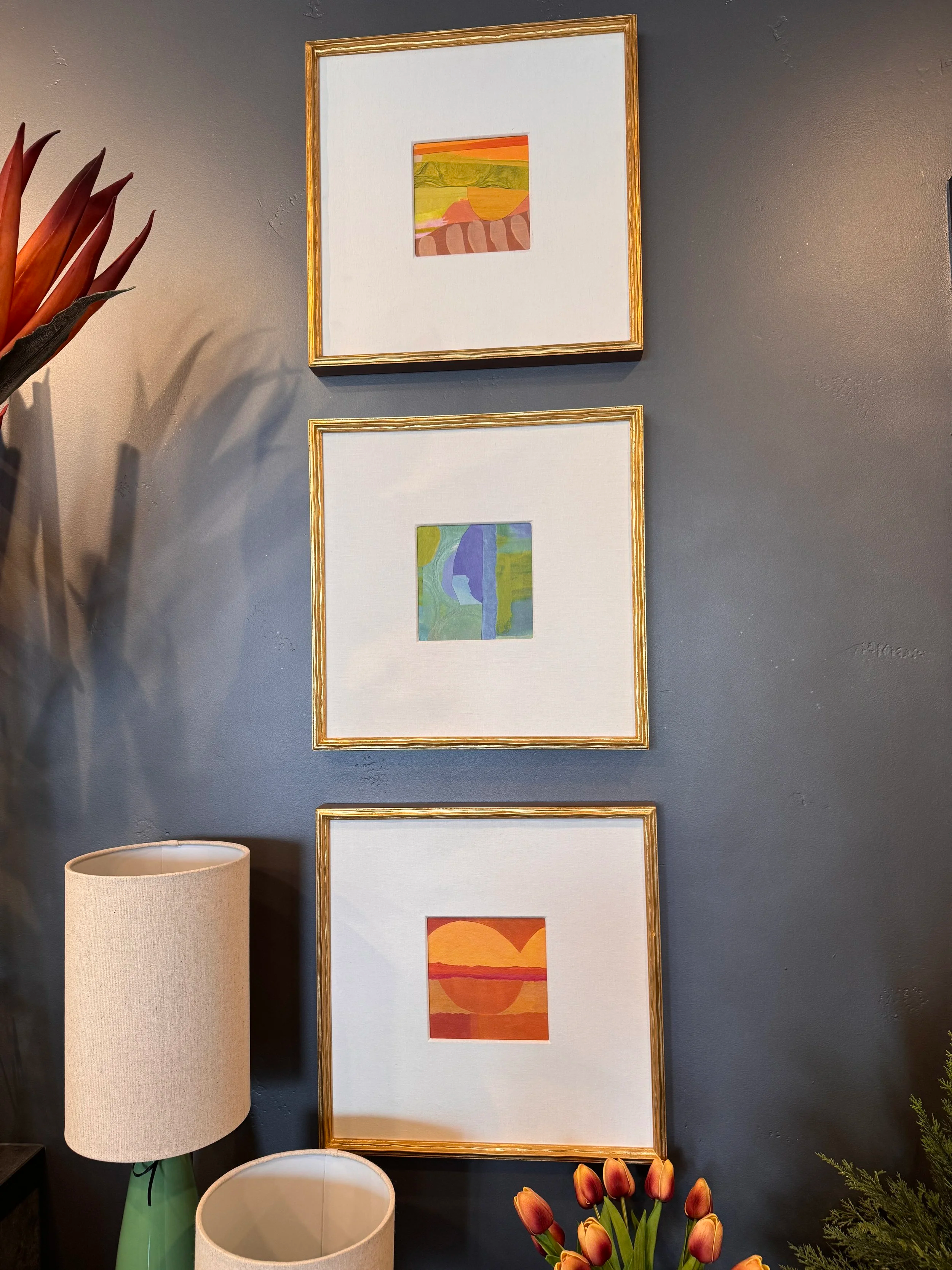 Decorative wall with three framed landscape paintings, a beige lamp with a green base, and a bouquet of orange tulips in a vase.