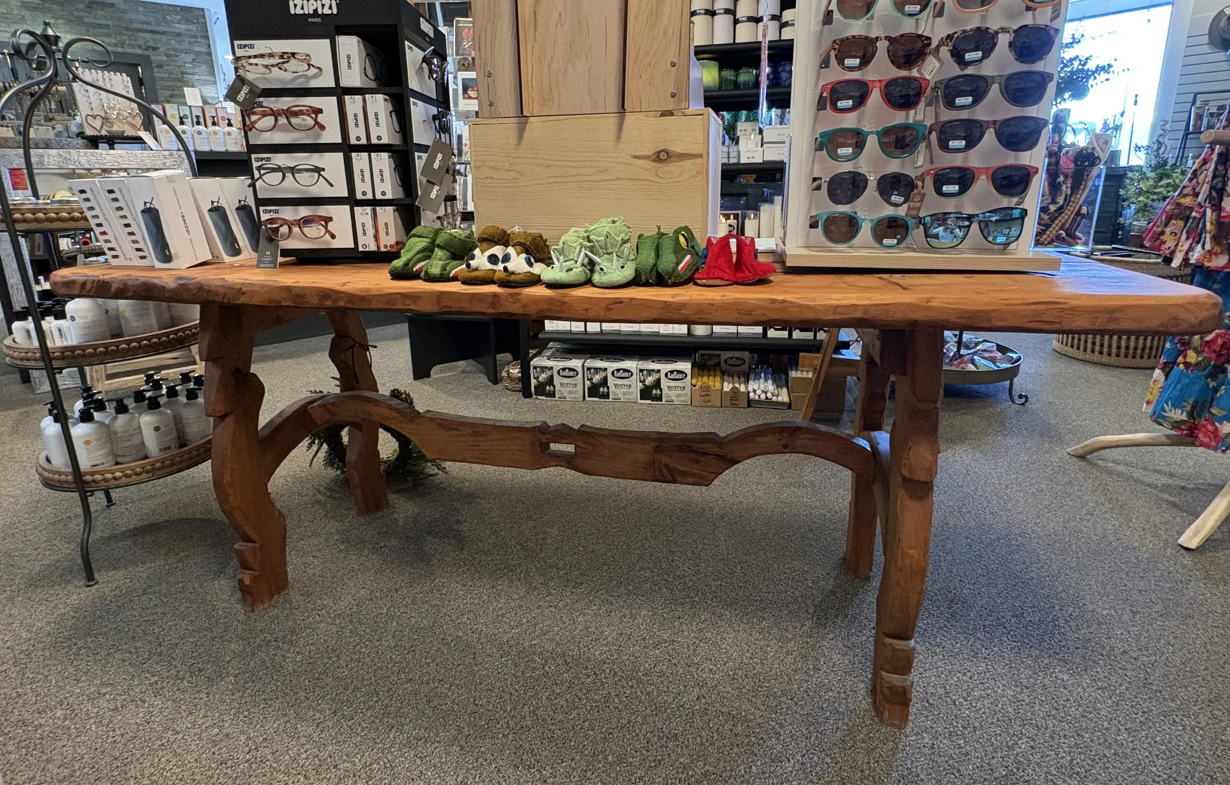 A rustic wooden table in a retail store displays small animal slippers, including frogs, bears, and a red ladybug. Behind the table are shelves with sunglasses and other accessories, with more eyewear and products visible in the background.