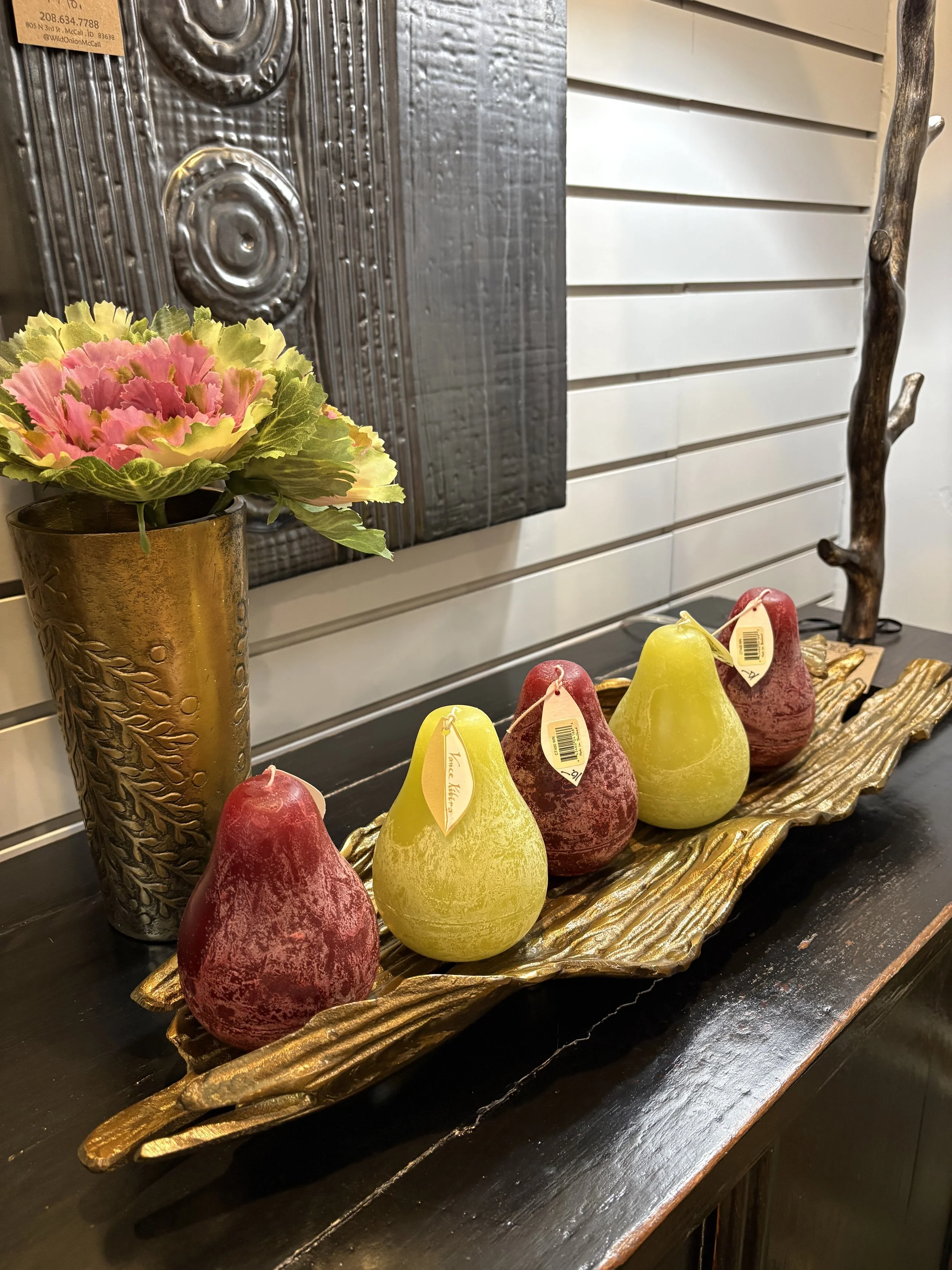 Decorative gold tray holding four artificial pears in red and yellow, a brass vase with a pink and yellow artificial flower, and a wooden sculptural branch, all on a black surface against a white paneled wall.
