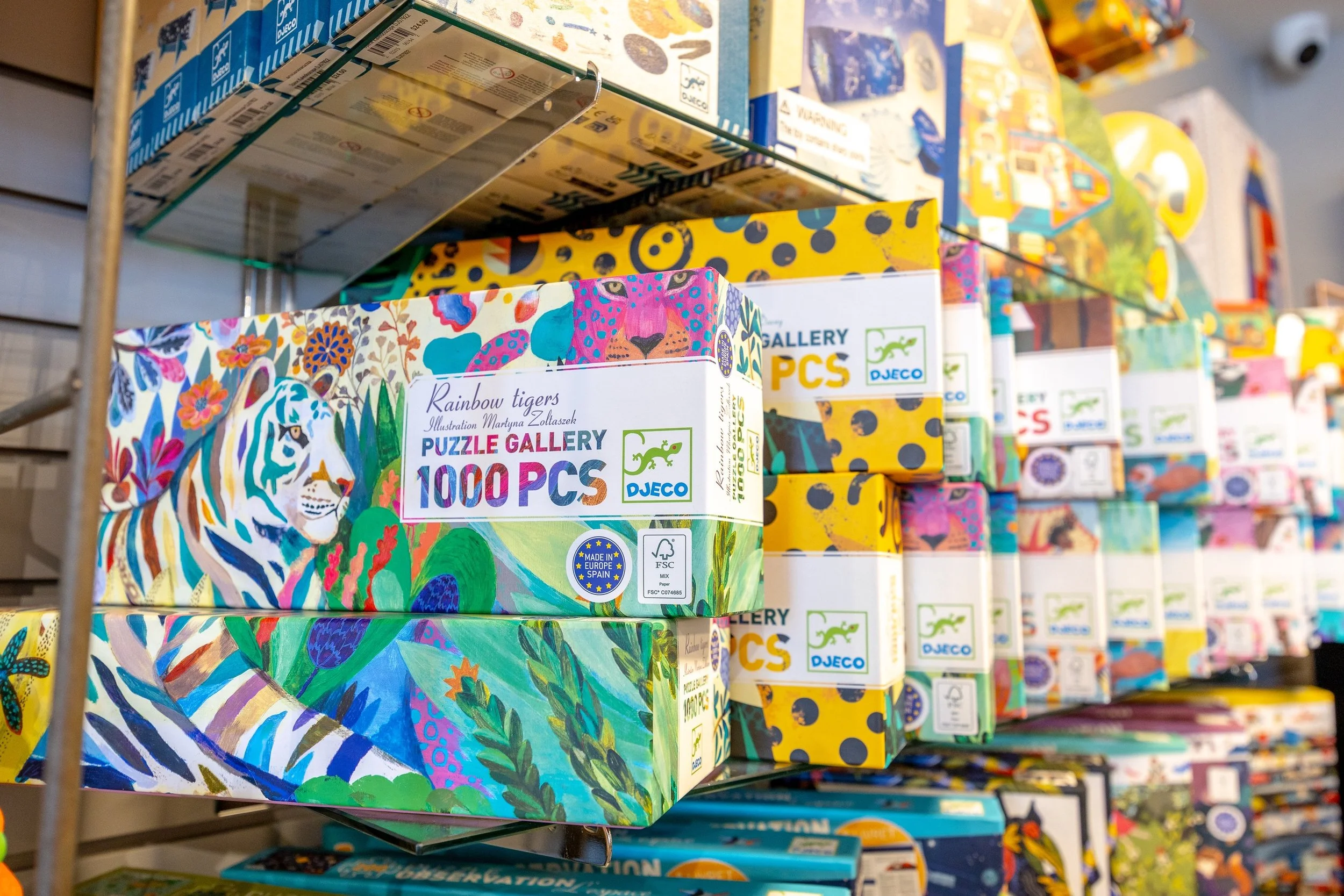 Colorful jigsaw puzzle boxes stacked on shelf, with the front box titled 'Rainbow Tigers' featuring a vibrant illustration of a tiger's face surrounded by floral and jungle patterns.