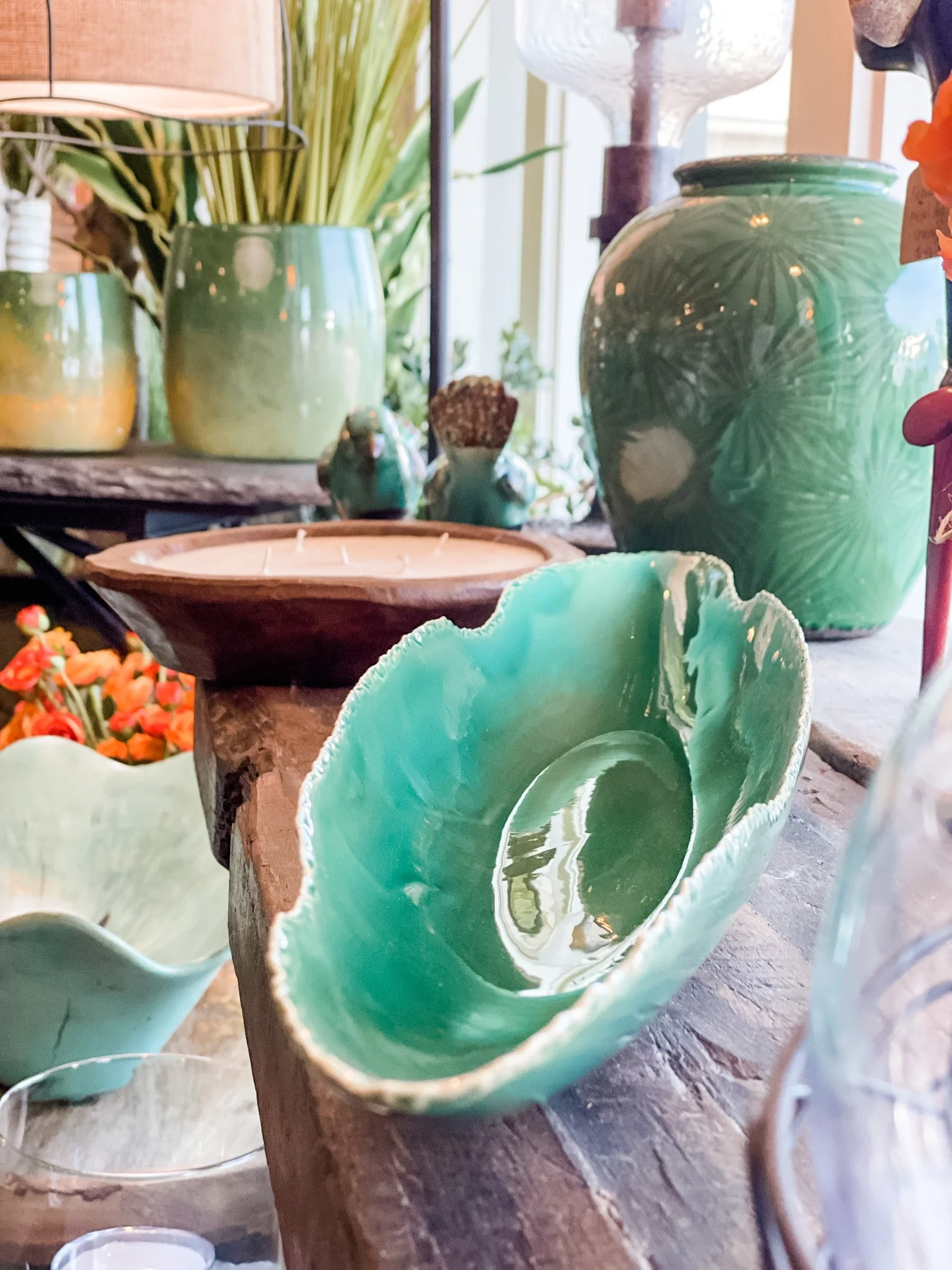 Close-up of green ceramic vases and bowls on a wooden surface, with plants and decorative items in the background.