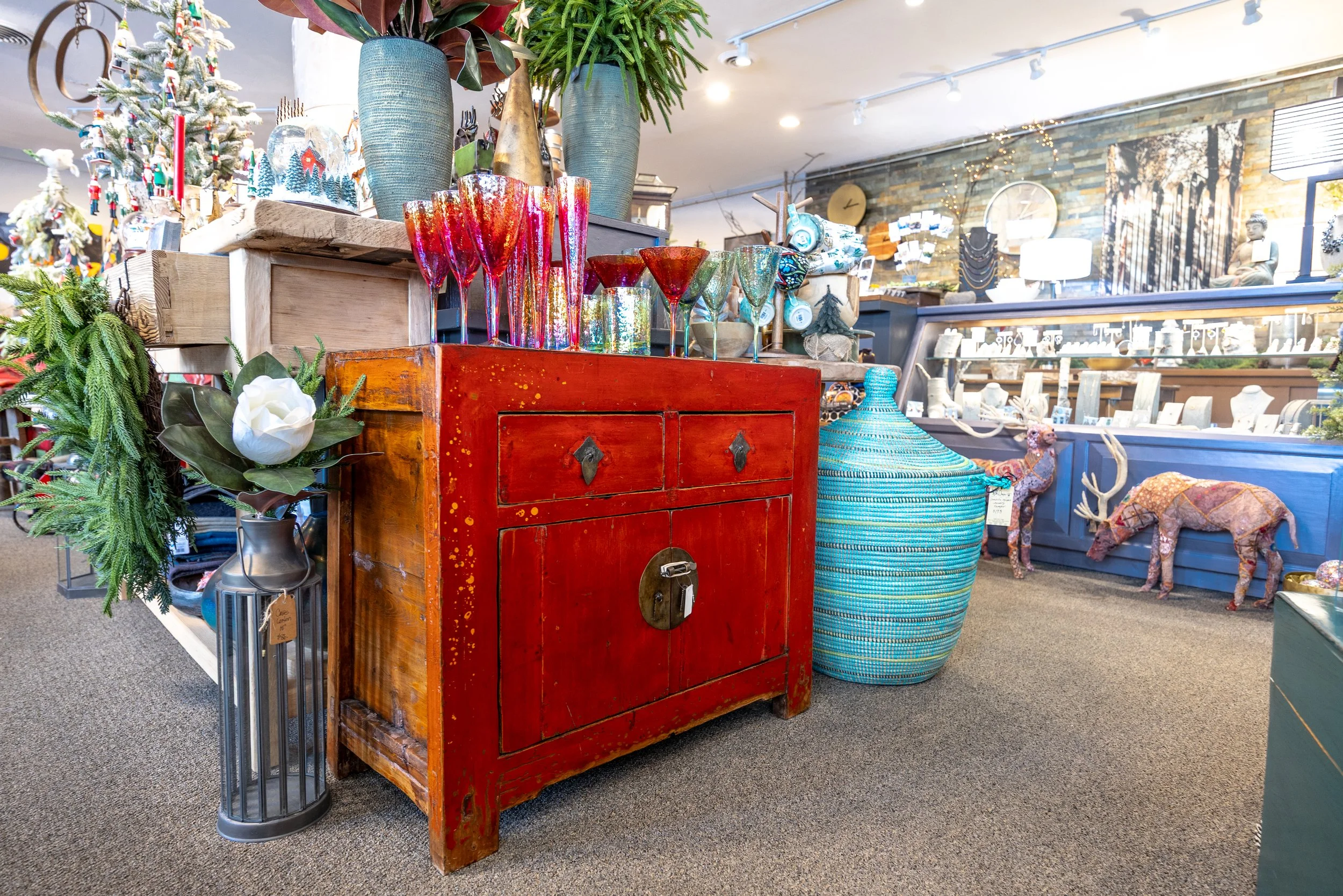Colorful glassware, potted plants, and decorative items on a wooden cabinet in a store with Christmas decorations and various home decor items in the background.