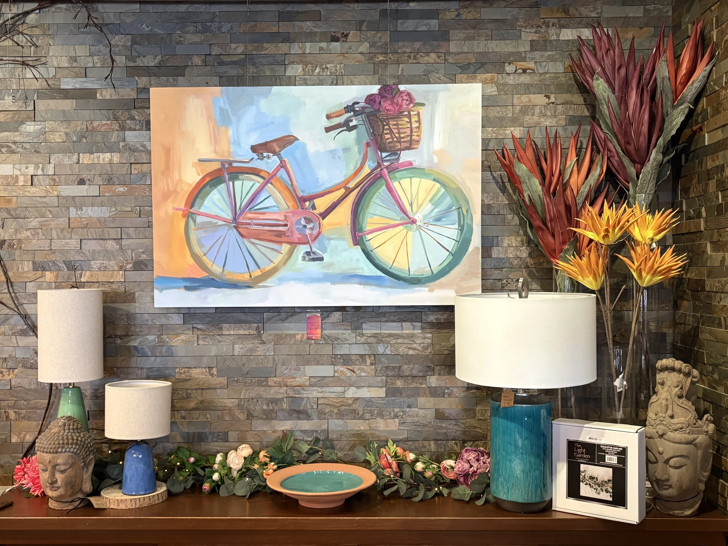 A colorful painting of a pink bicycle with a basket of pink flowers, hanging on a textured brick wall. Surrounding the painting are various decorative items including lamps, vases, and stone sculptures, with vibrant flowers in the foreground.
