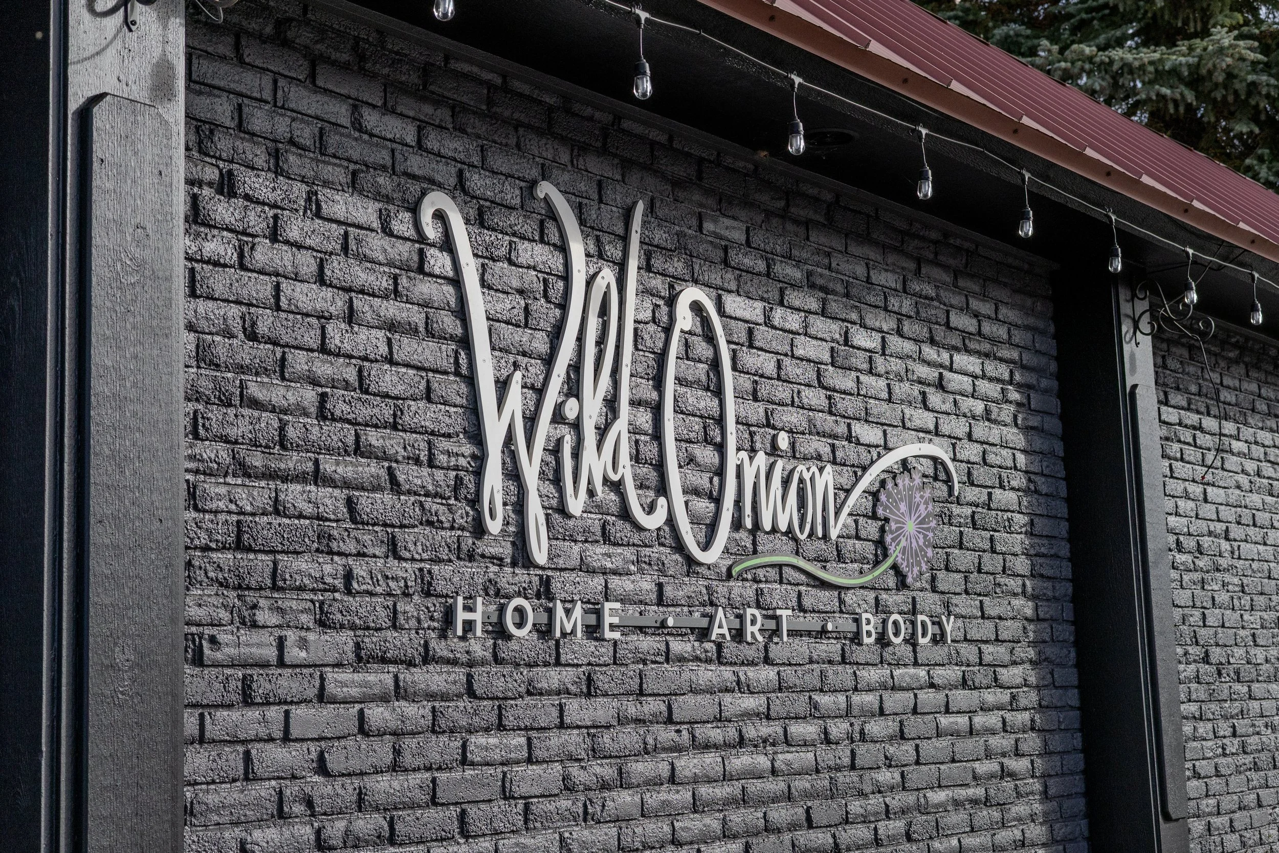 A black brick wall with metal lettering spelling 'Wild' and a pink decorative element, part of a sign for a home, art, and body business, with string lights hanging above.
