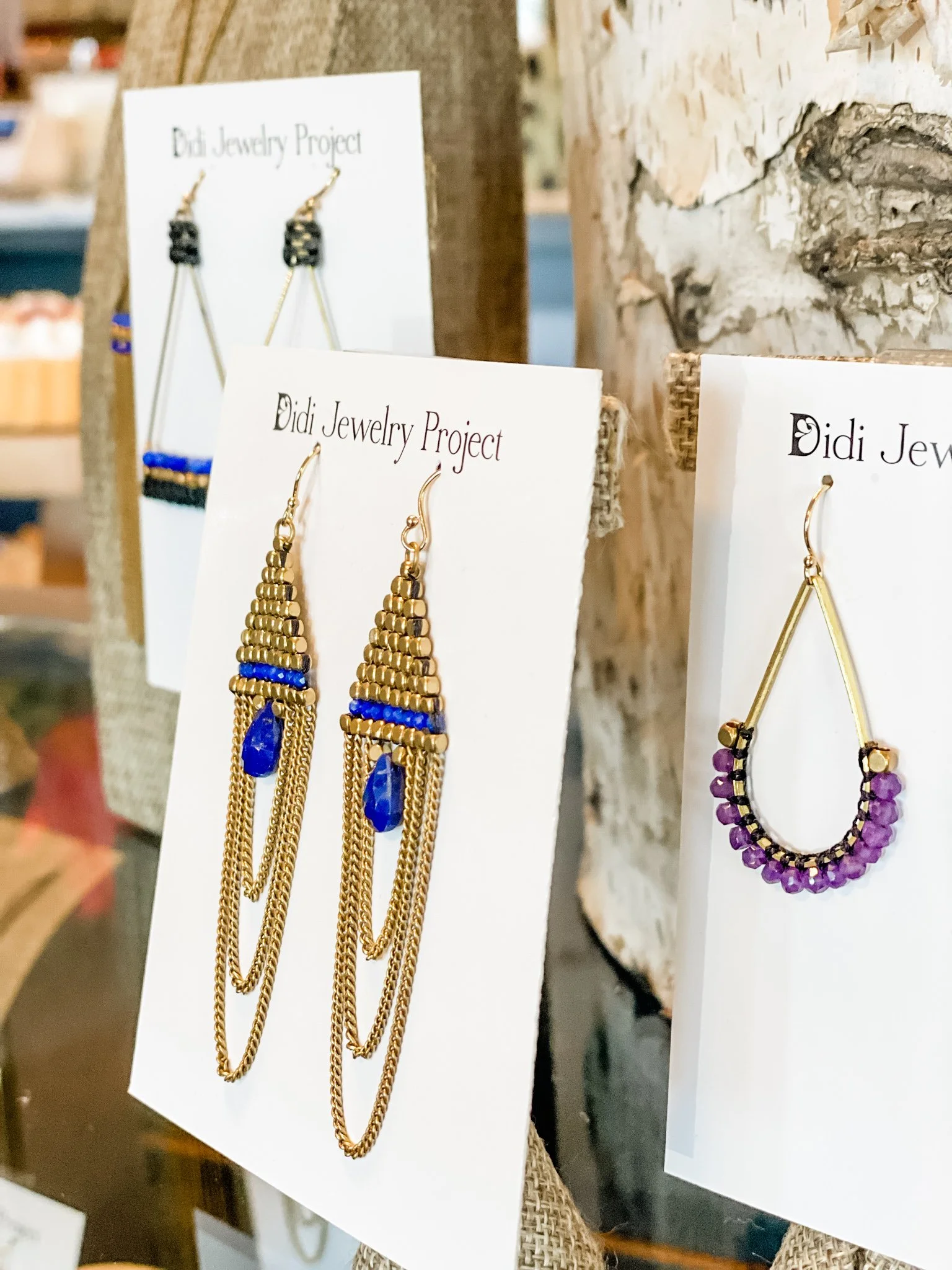 Two pairs of beaded earrings, one gold and blue, and the other gold and purple, displayed on white cards labeled "Didi Jewelry Project" at a craft fair or jewelry display.