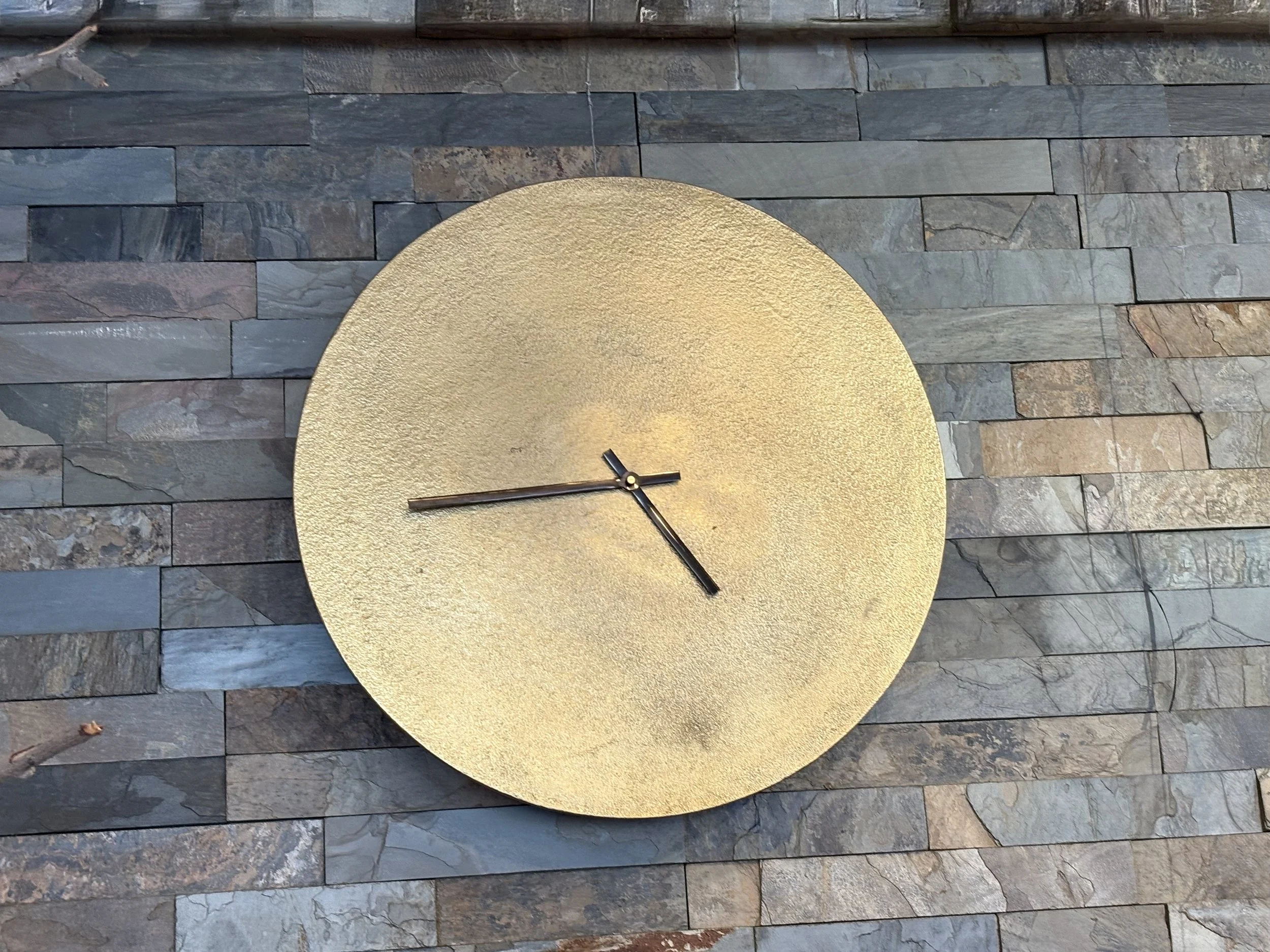 A gold-colored round clock with minimalistic black clock hands, mounted on a textured slate stone wall.