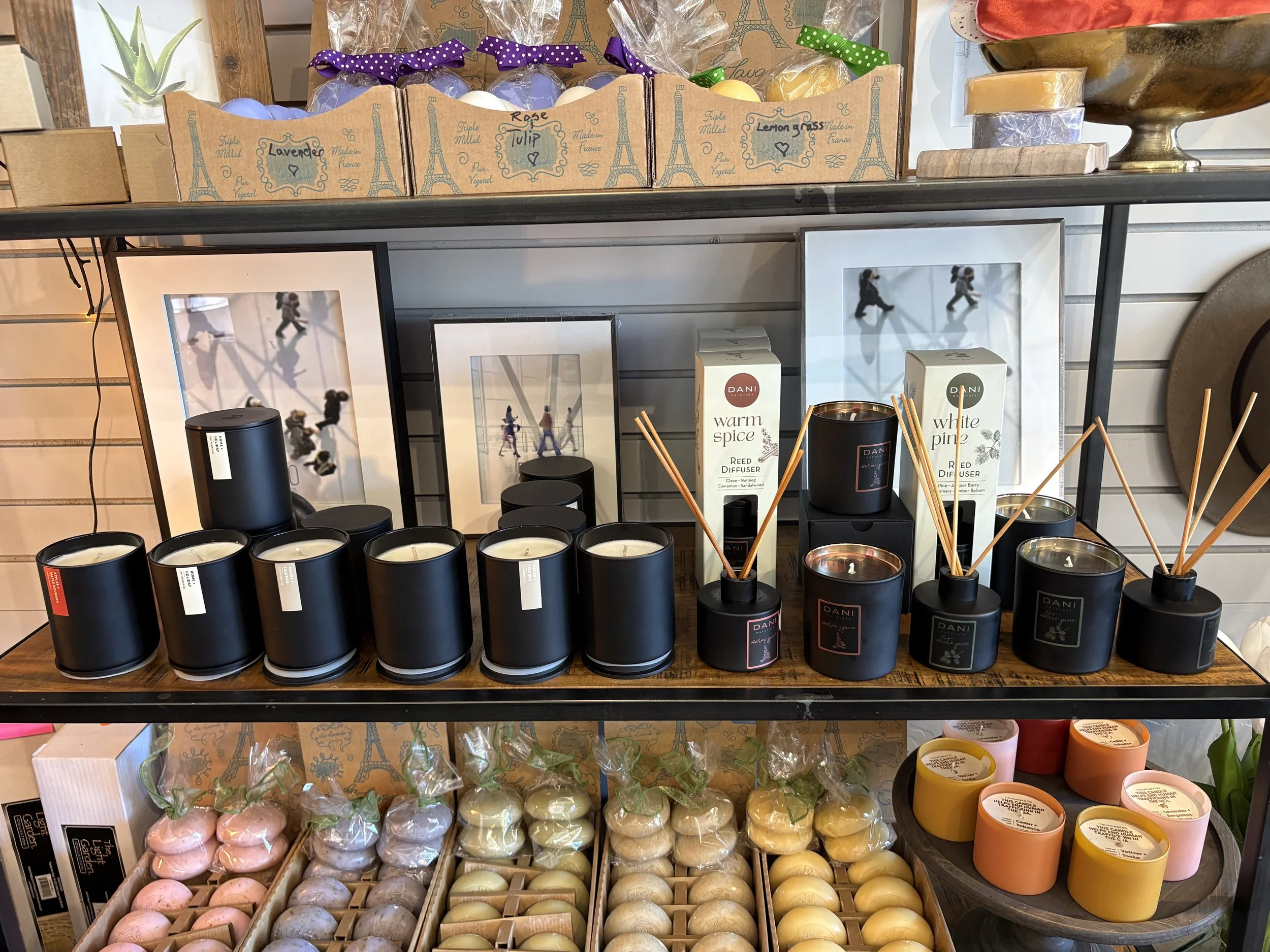 Shelf display of various scented candles in black, white, pink, yellow, cream, and green containers with reed diffusers and decorative bath bombs.