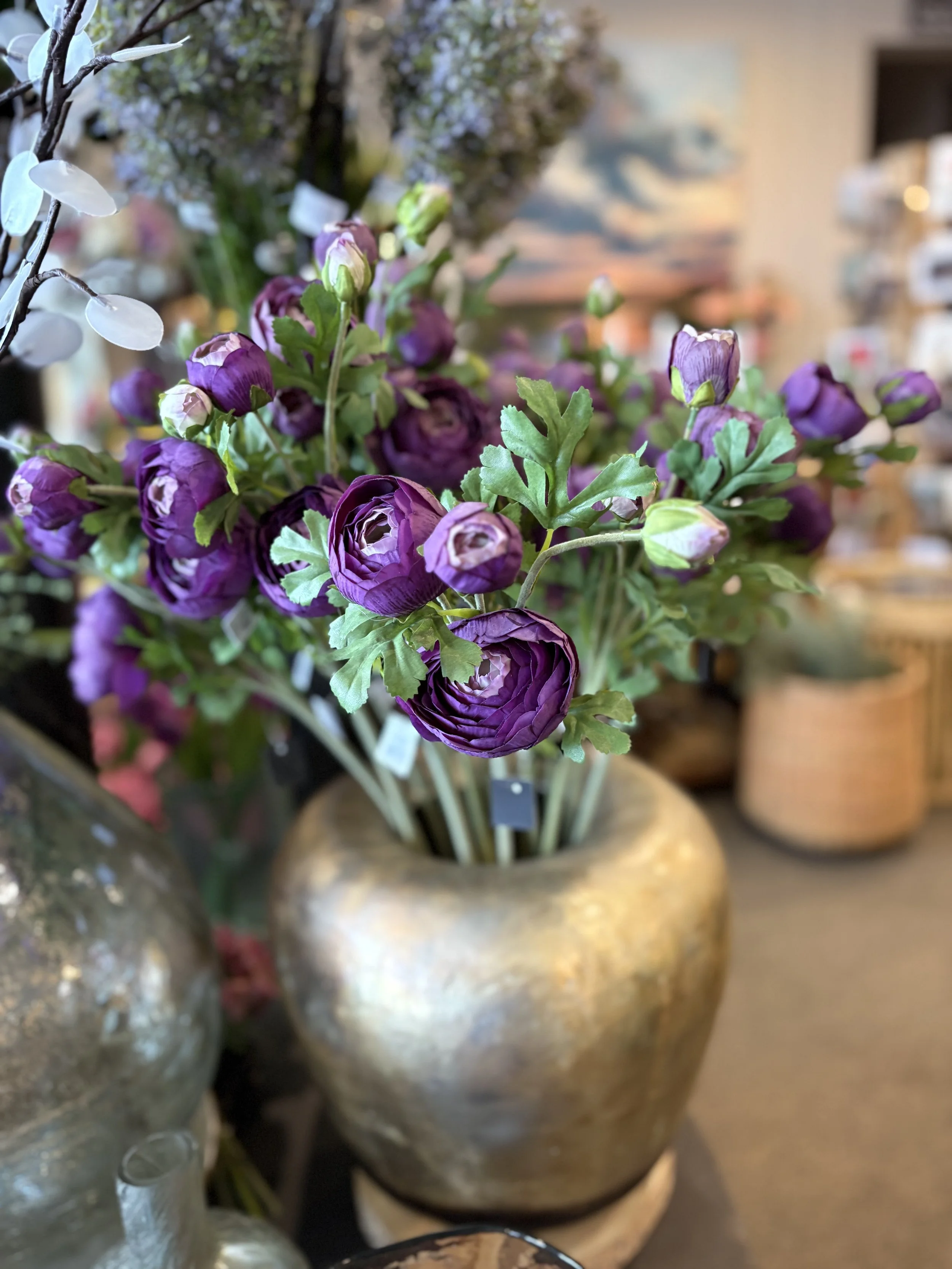 A close-up of a bouquet of purple and white ranunculus flowers in a gold-colored vase, with blurred background of other vases and floral arrangements.