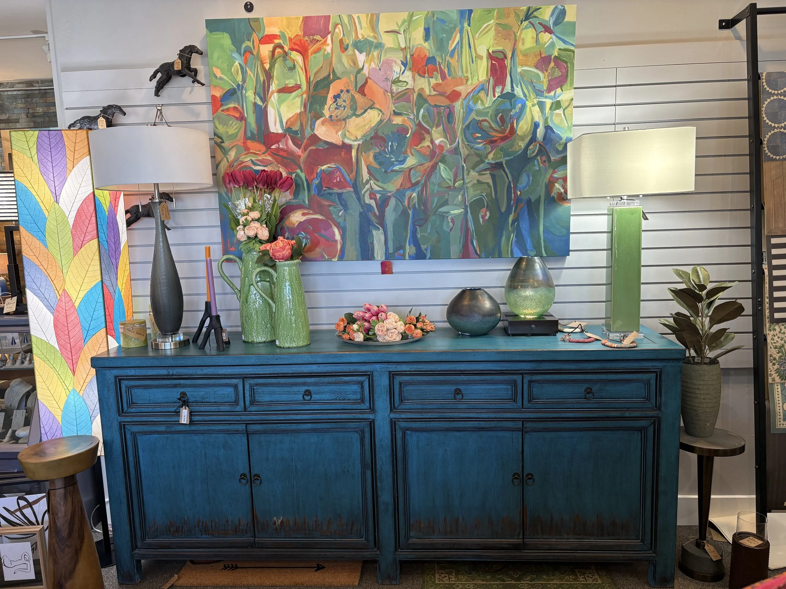 A collection of home decor items including a large abstract floral painting, green vases, a bouquet of pink and peach roses, two table lamps, and ceramic bowls on a blue antique-style sideboard.