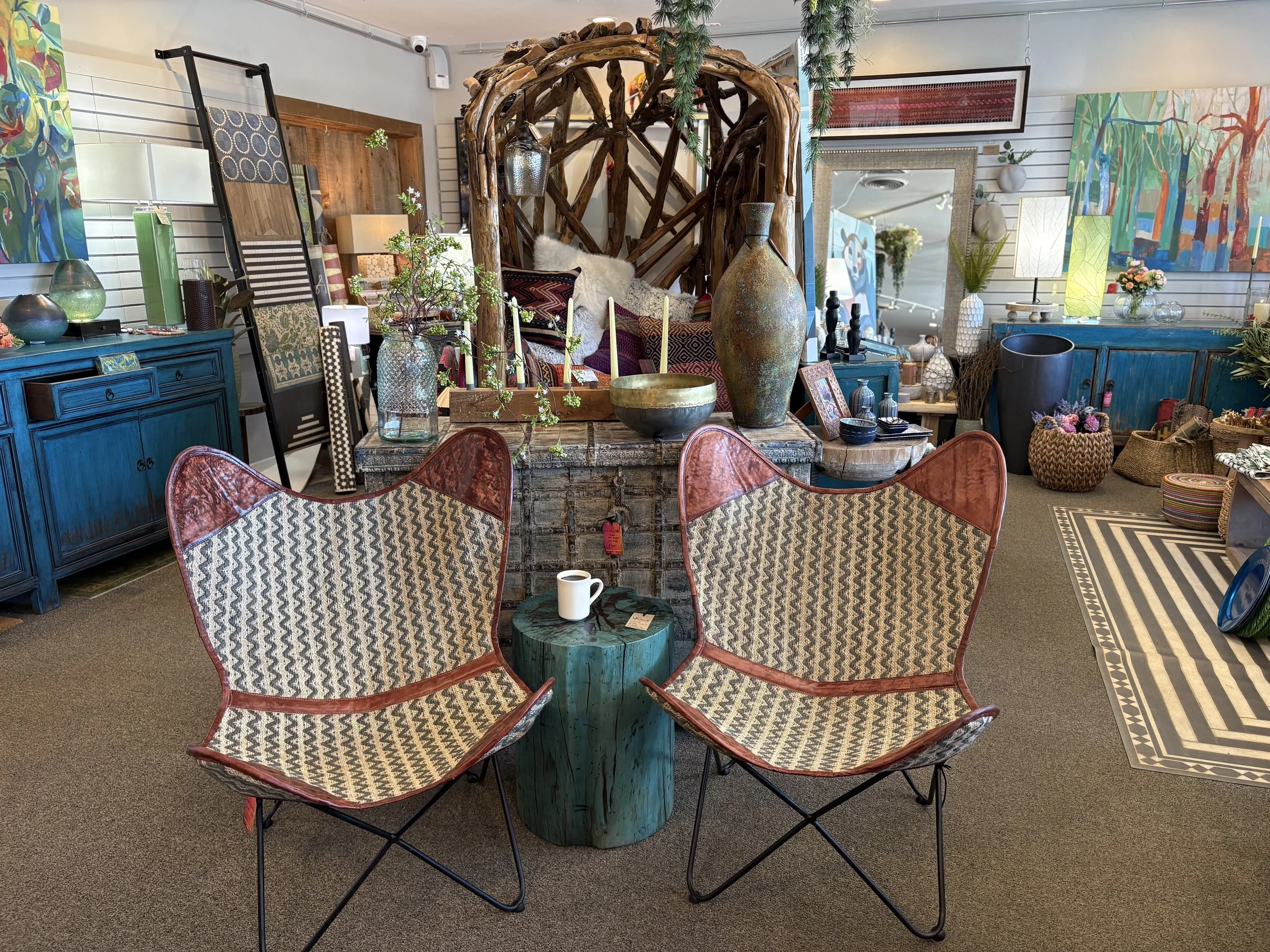 Display of decorative home interior items including patterned chairs, vases, artwork, baskets, and a rustic table with candles, in a cozy showroom setting.