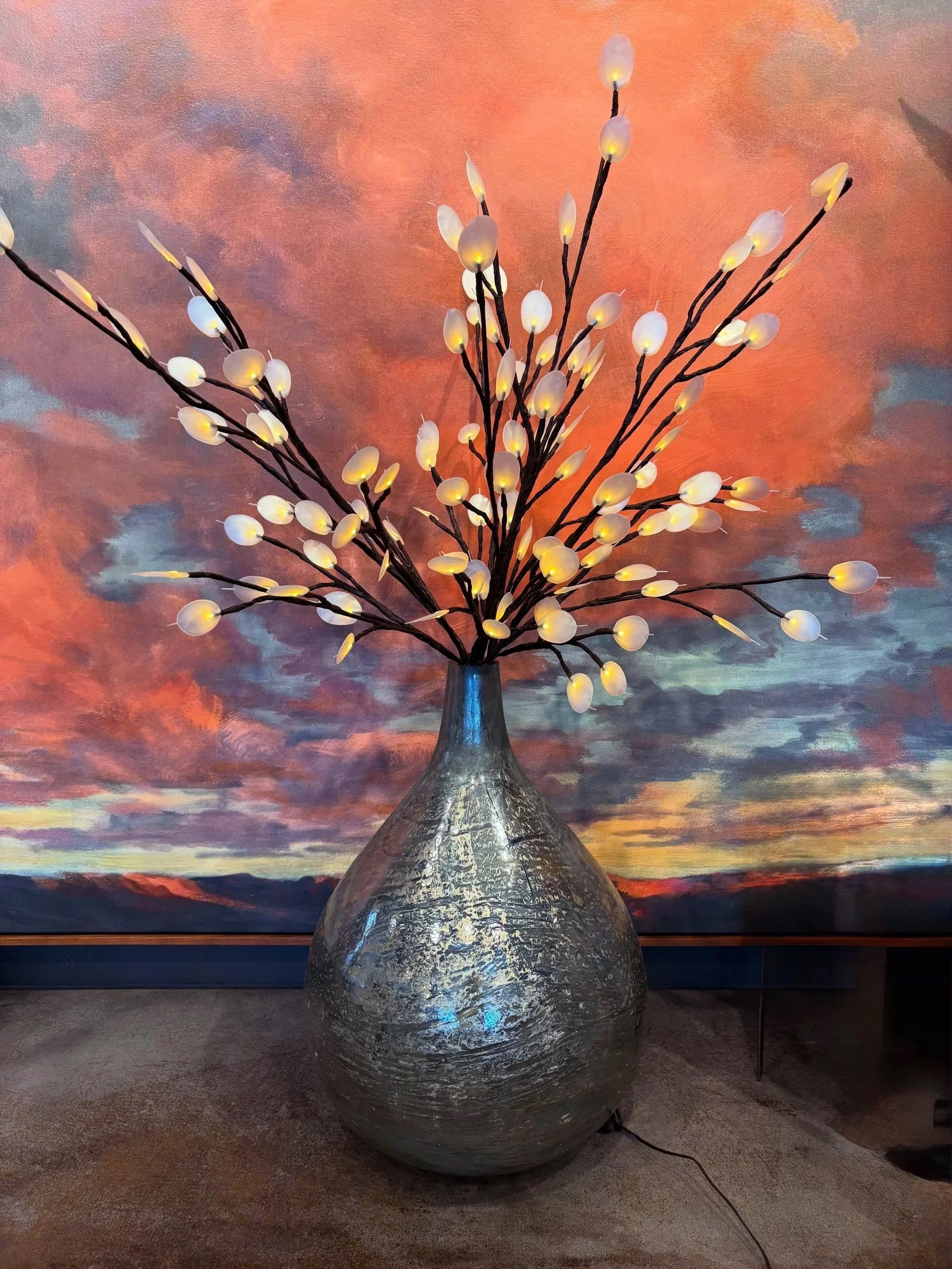 Large metallic vase with a textured surface holding a branch with numerous small white lights, against a colorful, abstract background with shades of pink, orange, purple, and blue.