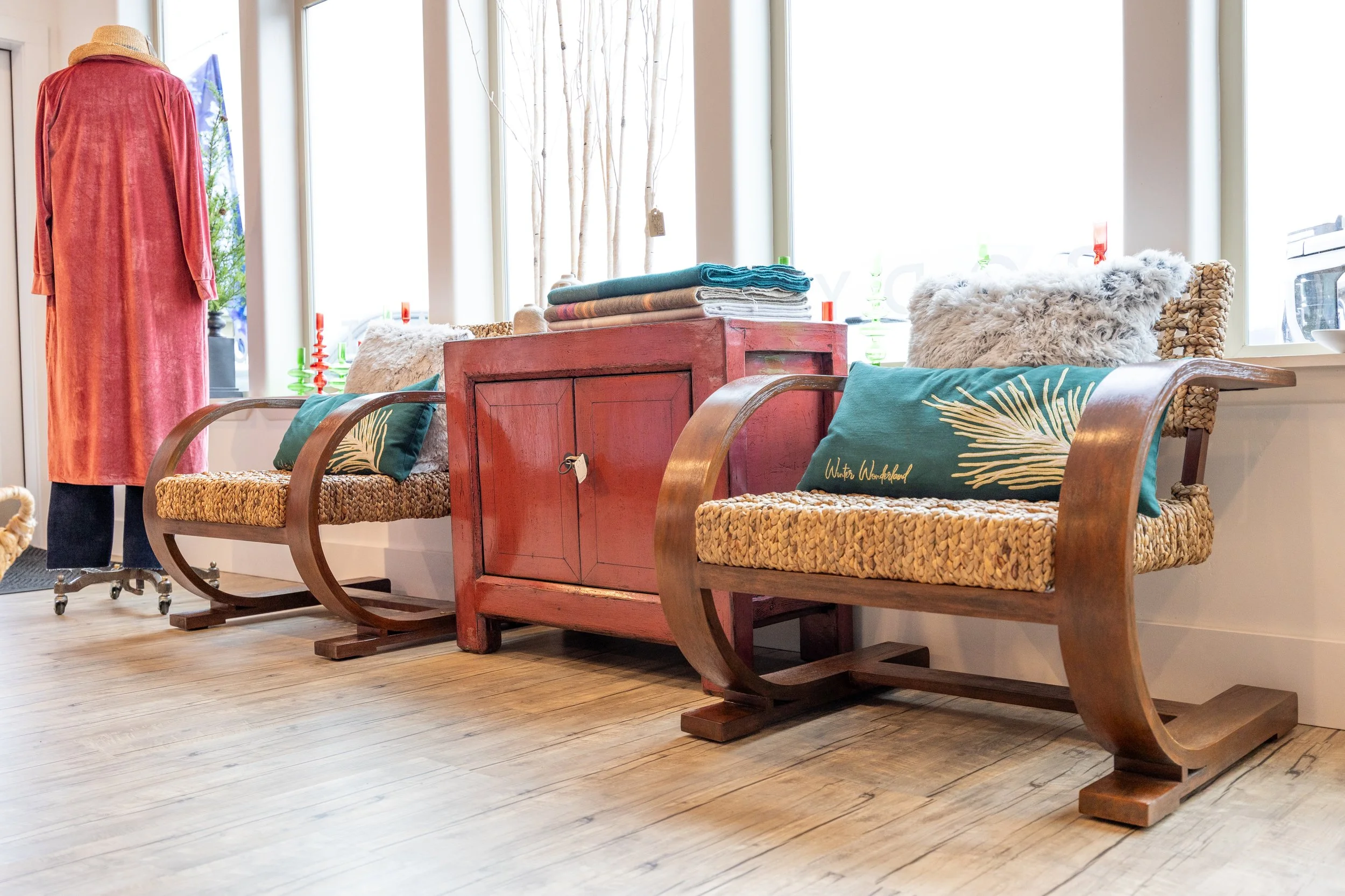 Interior of a store with wooden flooring, featuring two vintage wooden sleds used as display tables, decorated with pillows and folded blankets, near large windows with white frames.
