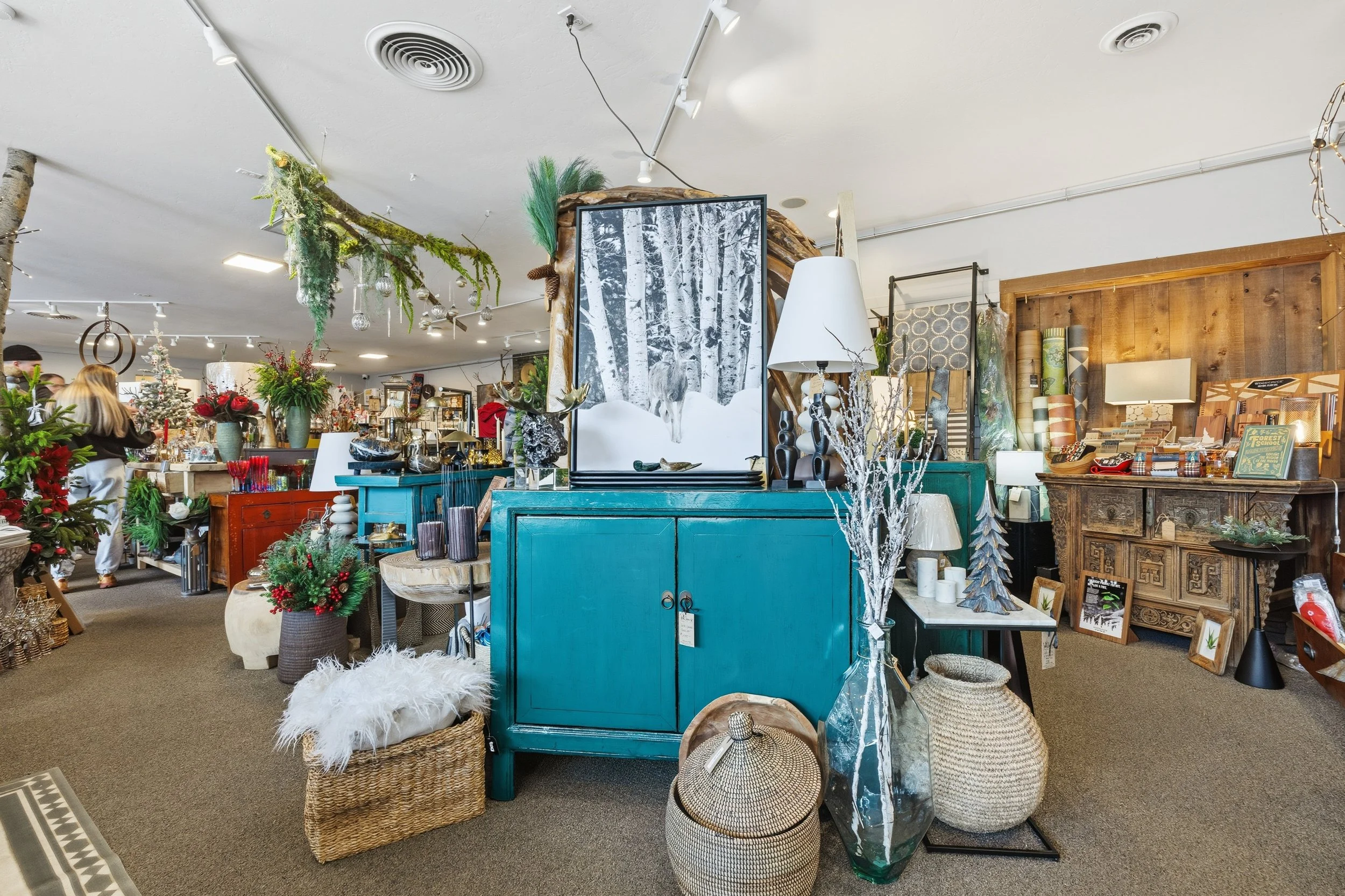 A store interior decorated with Christmas-themed items, plants, and home decor, including a prominent blue cabinet, pine branches, reindeer and winter scene artwork, wrapped baskets, and various decorative objects and prints.